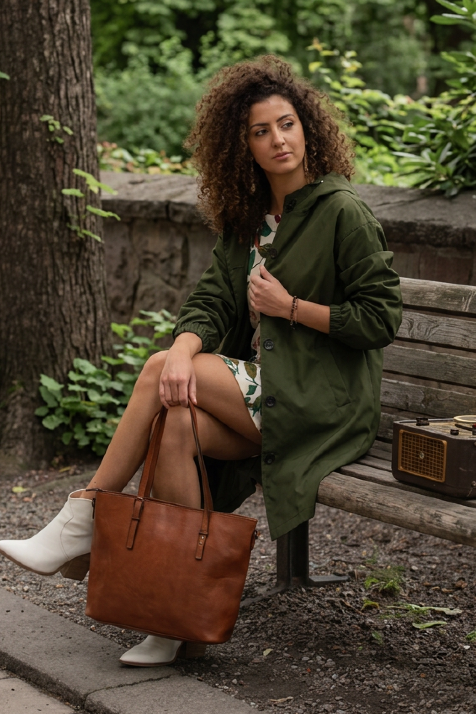 A woman with curly hair sits on a wooden bench in a park, wearing the Icon Windbreaker Rain Jacket, a patterned dress, and white boots. She holds a brown leather tote bag and looks to the side, with a vintage radio beside her.