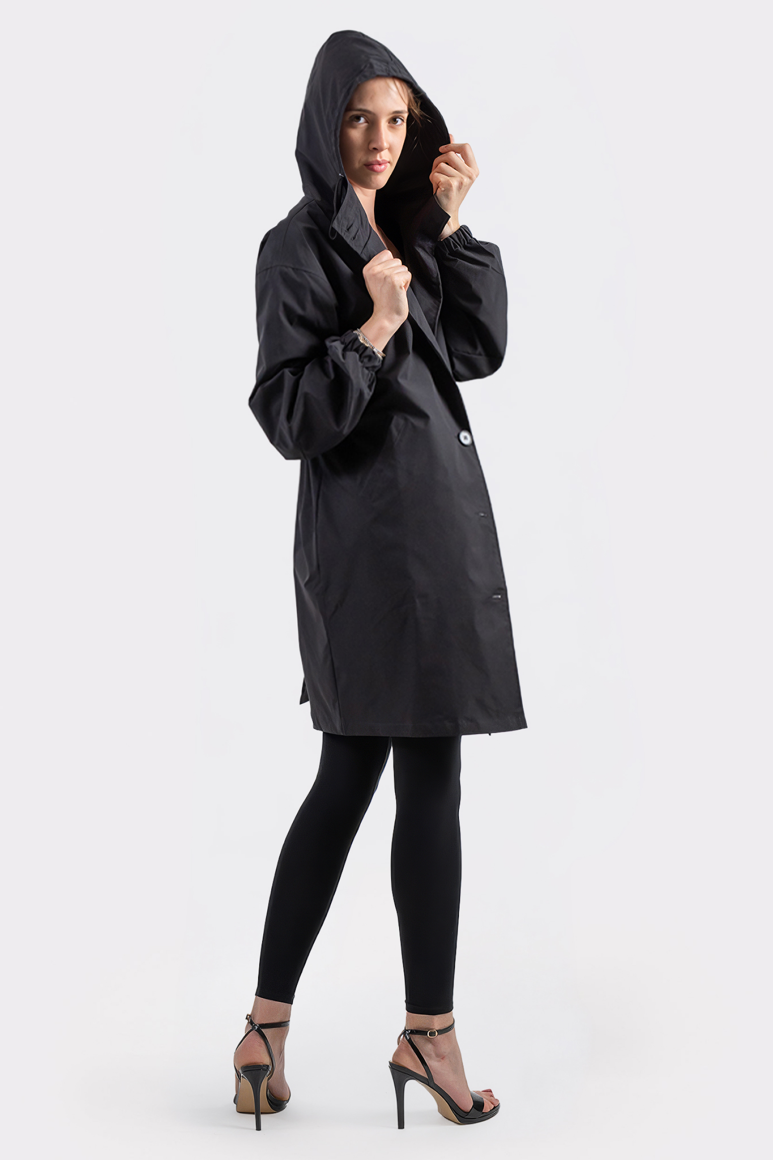 A person wearing the Icon Windbreaker Rain Jacket, black leggings, and high-heeled sandals stands against a plain white background, looking at the camera while holding the edge of the jacket’s hood with one hand.