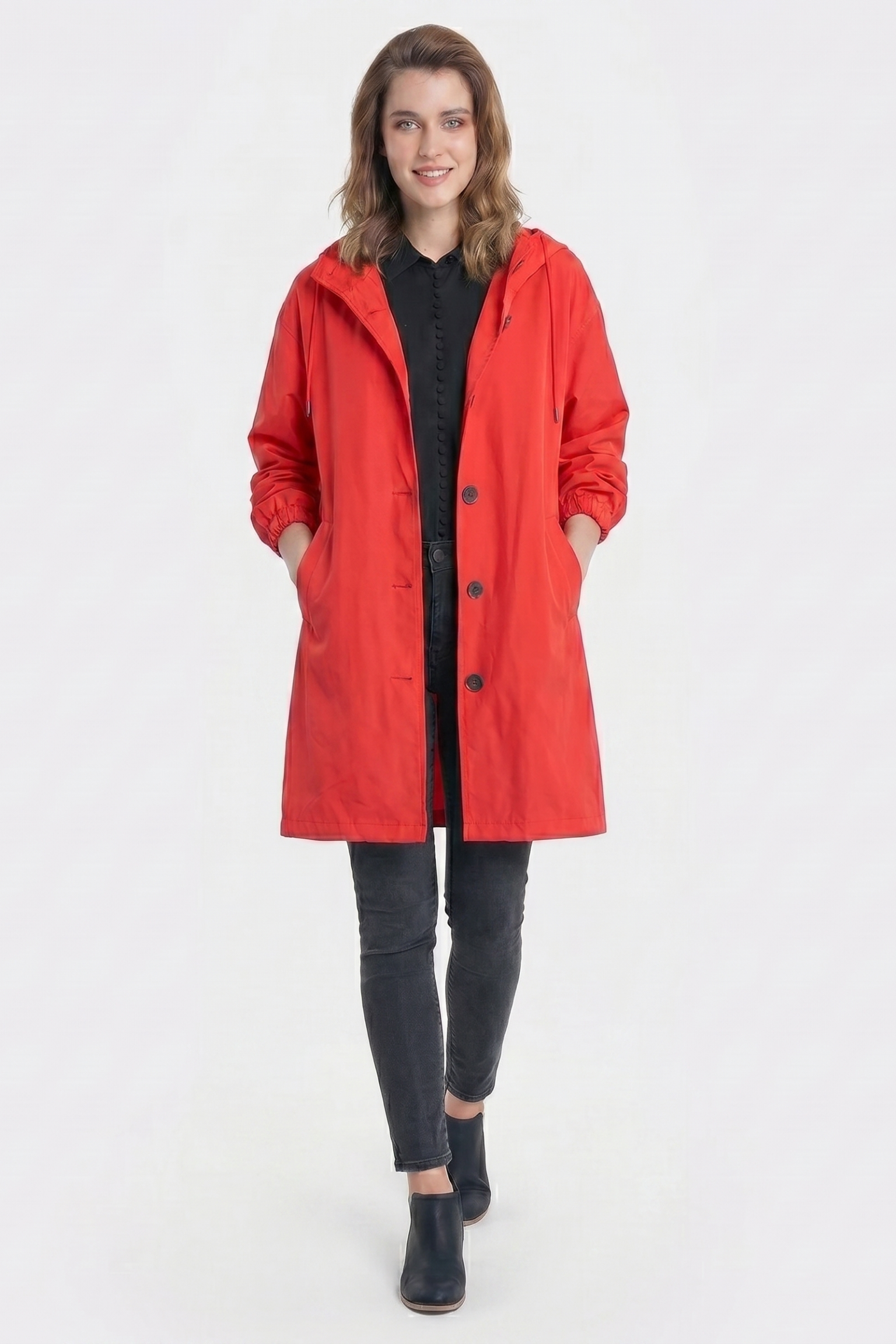 A person stands facing forward in the Icon Windbreaker Rain Jacket, bright red with a hood, over a black shirt and pants, hands in jacket pockets and wearing black shoes. The background is plain white.