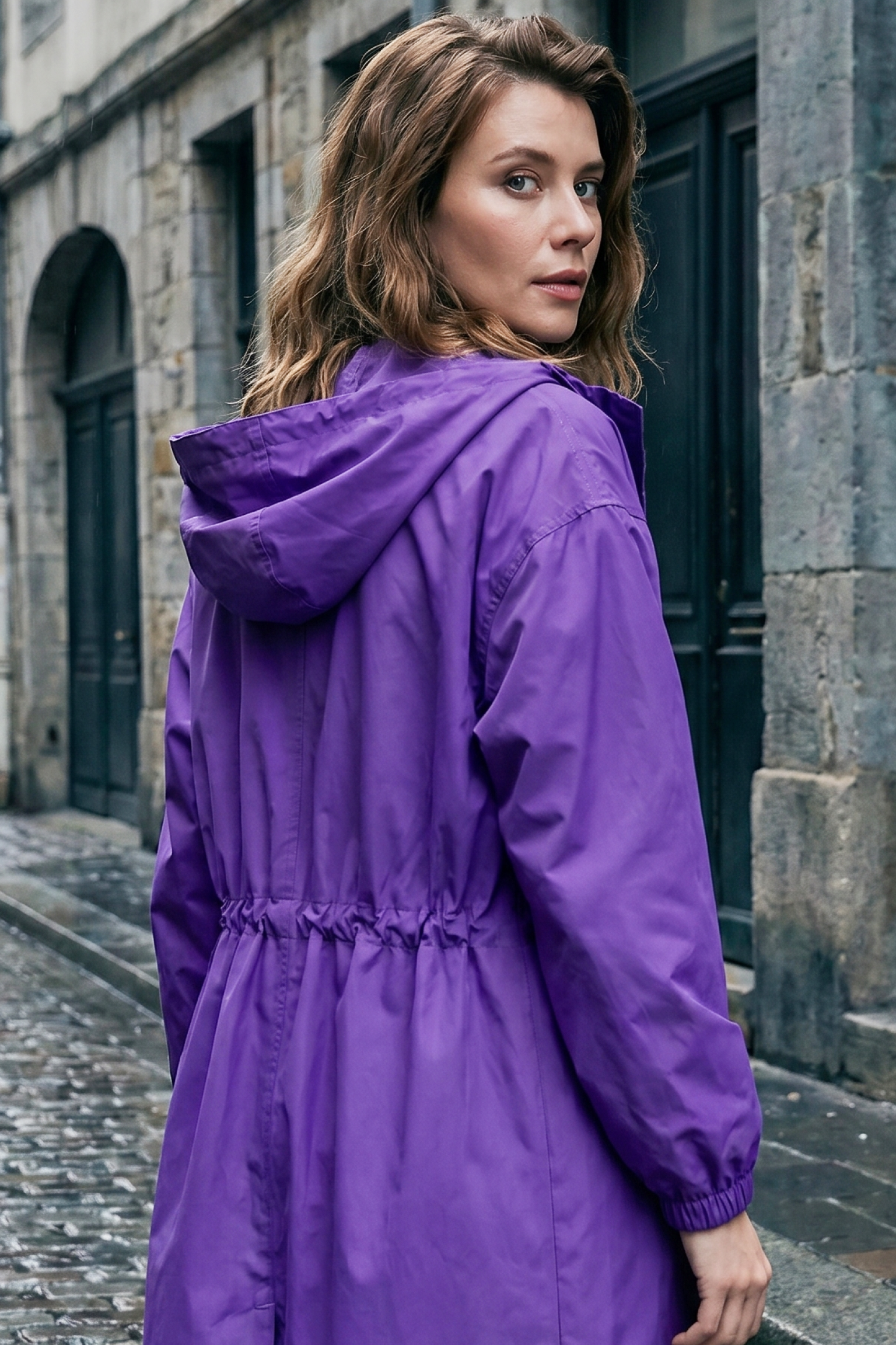 A woman with wavy brown hair stands on a cobblestone street, wearing the Icon Windbreaker Rain Jacket in purple. She looks over her shoulder toward the camera, with historic stone buildings in the background.