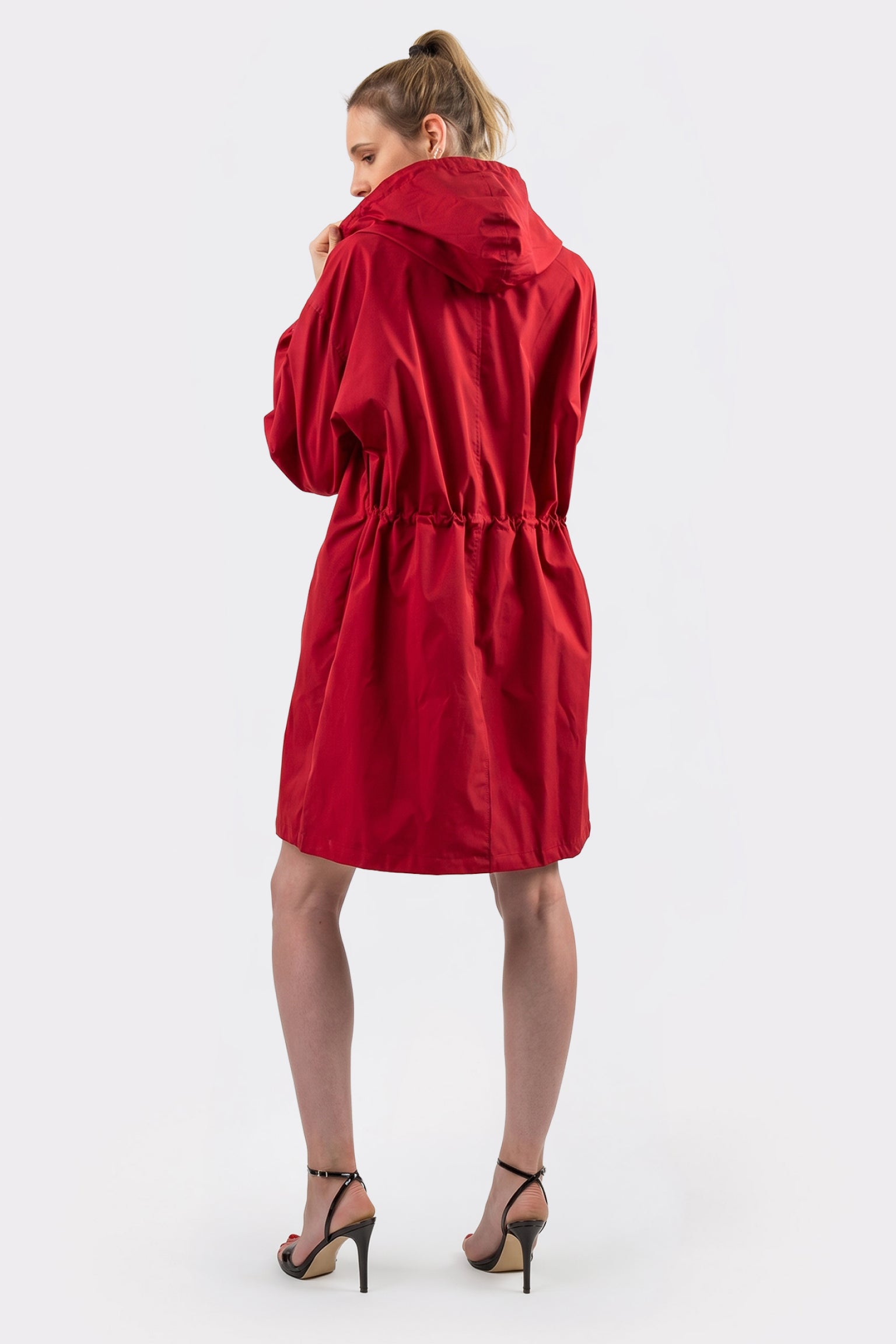 A person stands facing away, wearing the Icon Windbreaker Rain Jacket—a knee-length red hooded coat with an adjustable waist. They pair it with high-heeled, open-toe sandals against a plain light background.