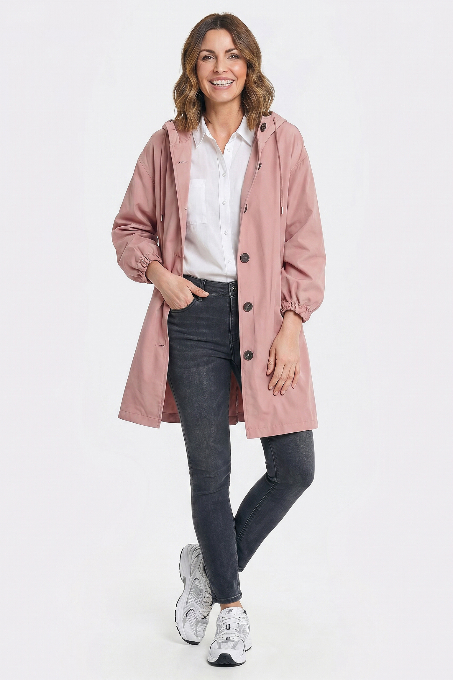 A woman smiles while wearing the Icon Windbreaker Rain Jacket in light pink, featuring a hood, paired with a white collared shirt, dark skinny jeans, and white sneakers. She stands against a plain white background with one hand in her pocket.