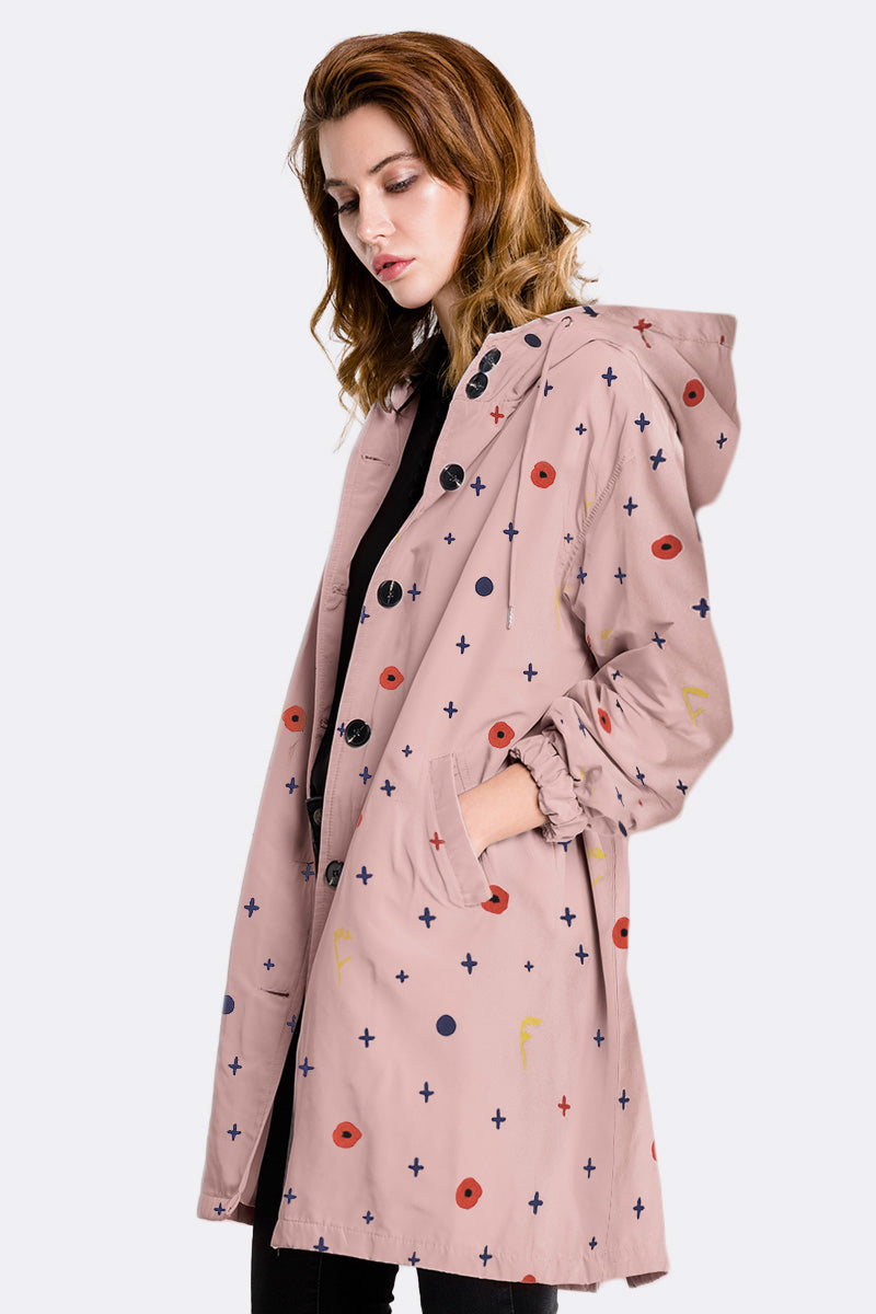 A woman with wavy light brown hair models the looselyboho DOT & BLOOM WINDBREAKER, a light pink water-resistant jacket with a geometric multicolored dot and cross print. She stands in profile, hand in pocket, against a plain light gray background.
