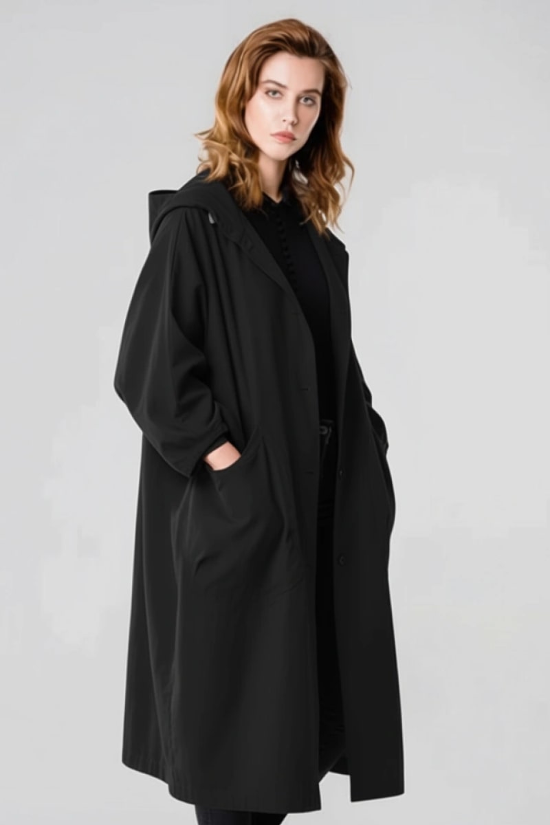 A woman with shoulder-length light brown hair stands against a plain background, wearing the looselyboho Black One Size Fits All Oversized Water Resistant Windbreaker Jacket. With her hands in the pockets, she gazes slightly to the side with a neutral expression.