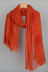 An orange *Color-POP Scarf* made of lightweight, textured fabric by *looselyboho* is draped over a wooden hanger against a neutral background. The all-weather scarf has frayed edges, adding a casual and cozy touch to its appearance.