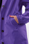 A person is wearing The Windbreaker Jacket's Gingham Ultraviolet Windbreaker. With black buttons and snug elastic cuffs, their left hand rests in the pocket, while bold orange fingernails add a pop of color to the look.