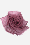 A mauve, textured scarf, crafted from ultra-breathable linen, is artfully arranged in a spiral pattern resembling a rose bloom. The scarf's frayed edges add to its artisanal charm. Set against a plain white background, the rich color and intricate details of this versatile *Lightweight Linen Scarf* by *looselyboho* are highlighted beautifully.