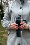 A person wearing a gray raincoat holds a dark blue, vacuum-insulated Indestructible Travel Cup by looselyboho with a straw and handle in both hands. The background shows green grass, trees, and an overcast sky. The person's nails are painted pink, and they are wearing a ring on their left hand.