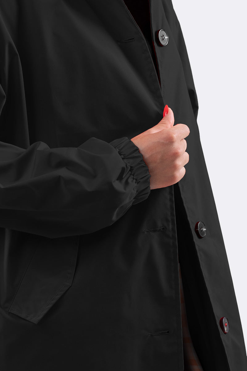 A person stands against a plain background, wearing the LUXE BLACK WATERPROOF WINDBREAKER by looselyboho. Their red-polished nails grasp the edge near buttons of the water-resistant coat, which includes side pockets, elastic cuffs, and an adjustable hood for versatility.