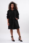 Against a white background, a person with curly hair wears the LUXE BLACK WATERPROOF WINDBREAKER by looselyboho. The sleek, water-resistant coat has an adjustable hood and complements their black high heels as they look slightly to the side with one hand in their pocket.