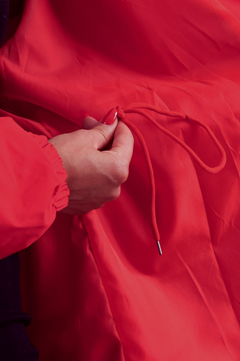 A person is adjusting the fit of a red Water Resistant Oversized Hooded Windbreaker Rain Jacket by pulling its matching drawstring. The person's hand is partially visible, highlighting the timeless style and versatility of The WJacket.