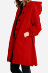 A person wearing a long, red Water Resistant Oversized Hooded Windbreaker Rain Jacket from The WJacket, complete with black buttons and a hood, stands against a plain white background. The jacket, showcasing timeless style, features pockets where the person has one hand tucked in. They are also dressed in black pants.