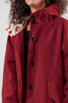 A person in a hooded red coat, similar to the looselyboho BURGUNDY BLOSSOM WINDBREAKER, faces forward. The coat has black buttons and a floral-patterned lining. Their curly hair enhances the hoop earrings they're wearing. Only their upper body is visible.