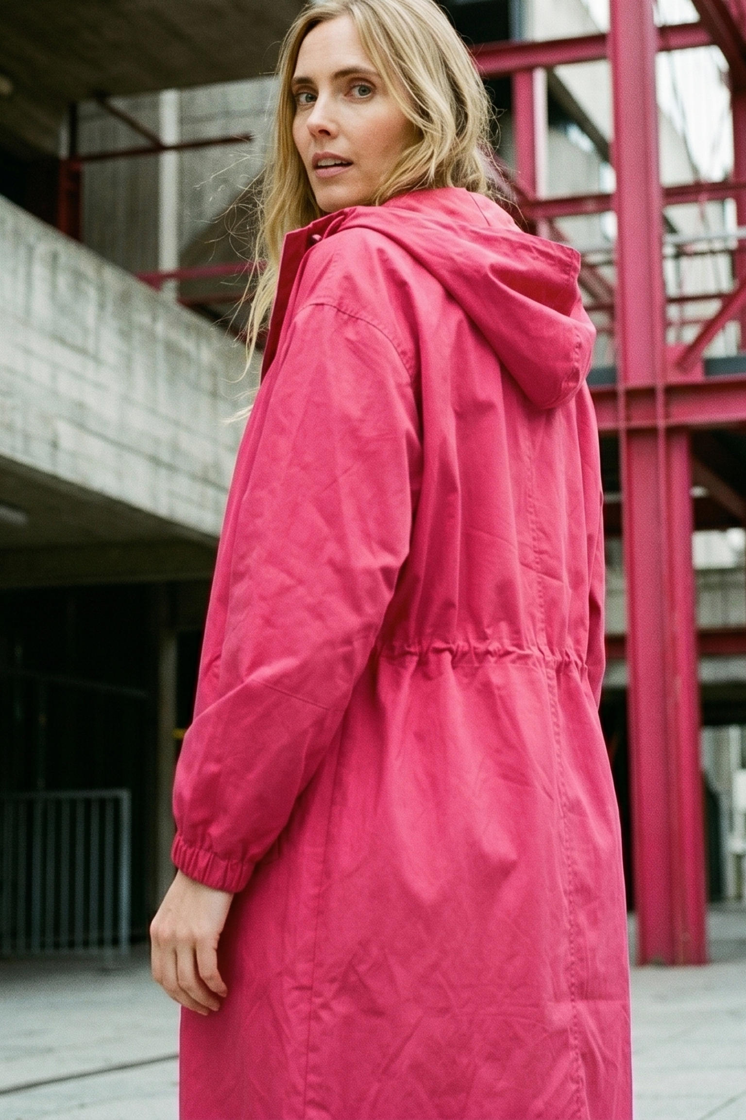 A person with long blonde hair is outdoors, wearing The Water Resistant Hooded Oversized Windbreaker Rain Jacket in bright pink. Concrete and red metal structures are in the background as they look over their shoulder at the camera.