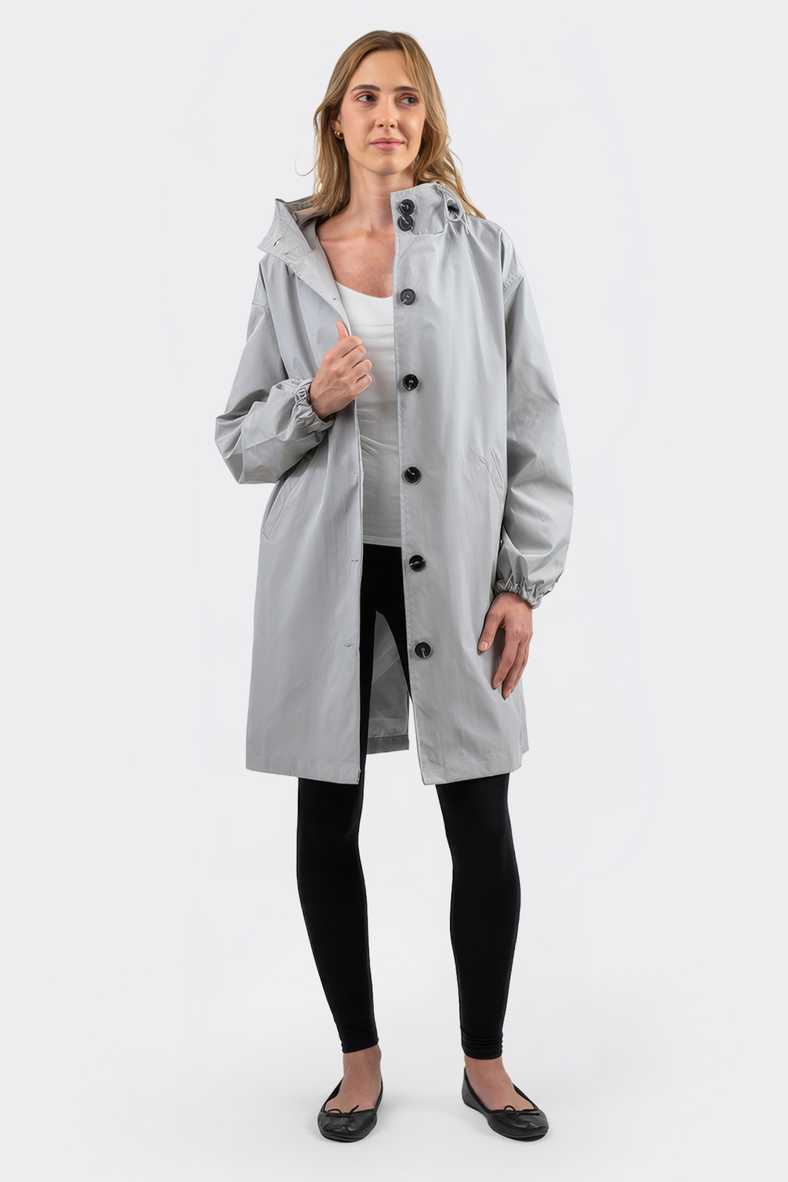 A person stands facing forward, wearing the Icon Windbreaker Rain Jacket in light gray with black buttons, paired with black leggings and flats. The background is plain white.