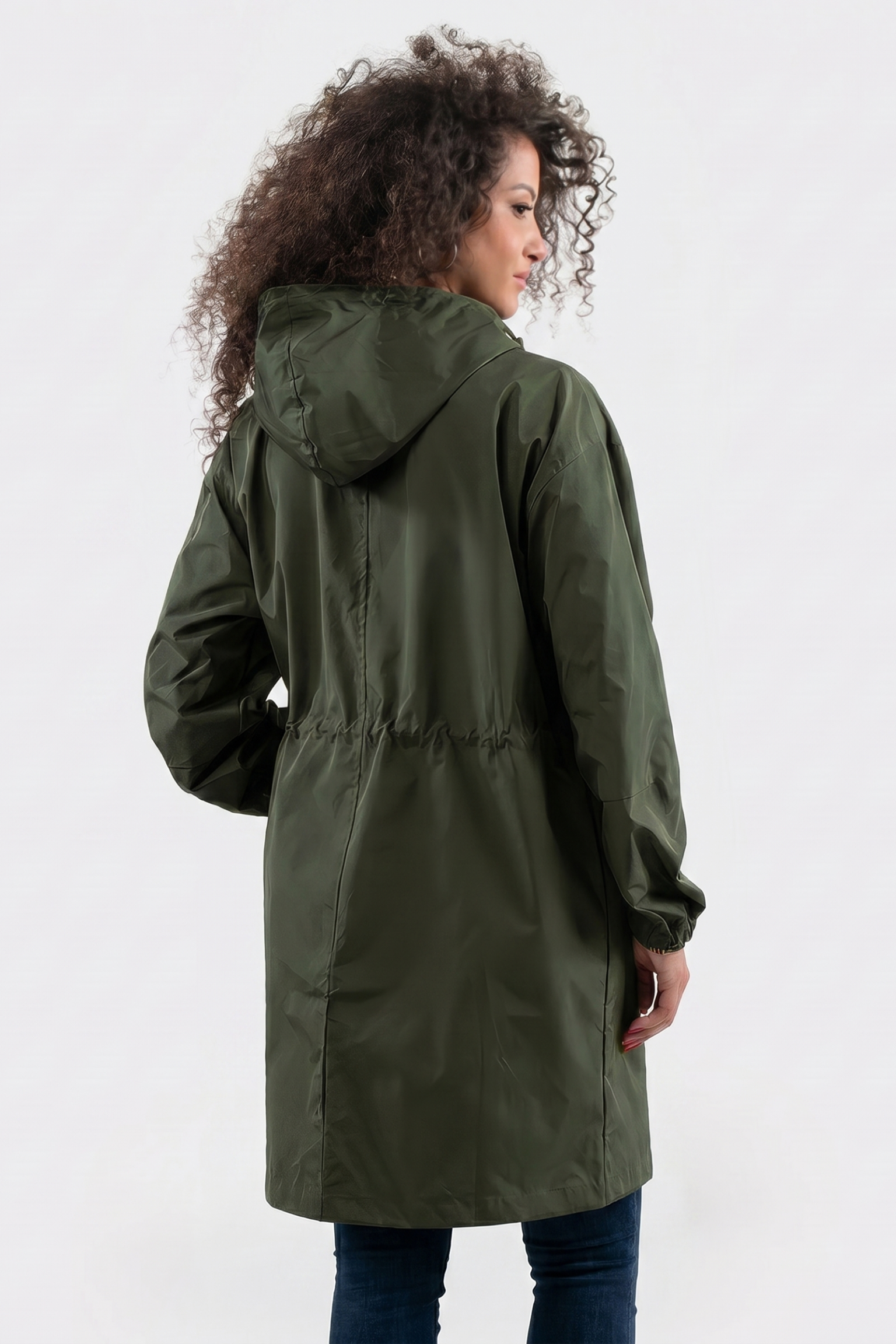 A person with curly hair, wearing dark blue jeans, stands facing away to show the back of the Icon Windbreaker Rain Jacket—a long, olive green hooded coat with long sleeves and a relaxed fit.