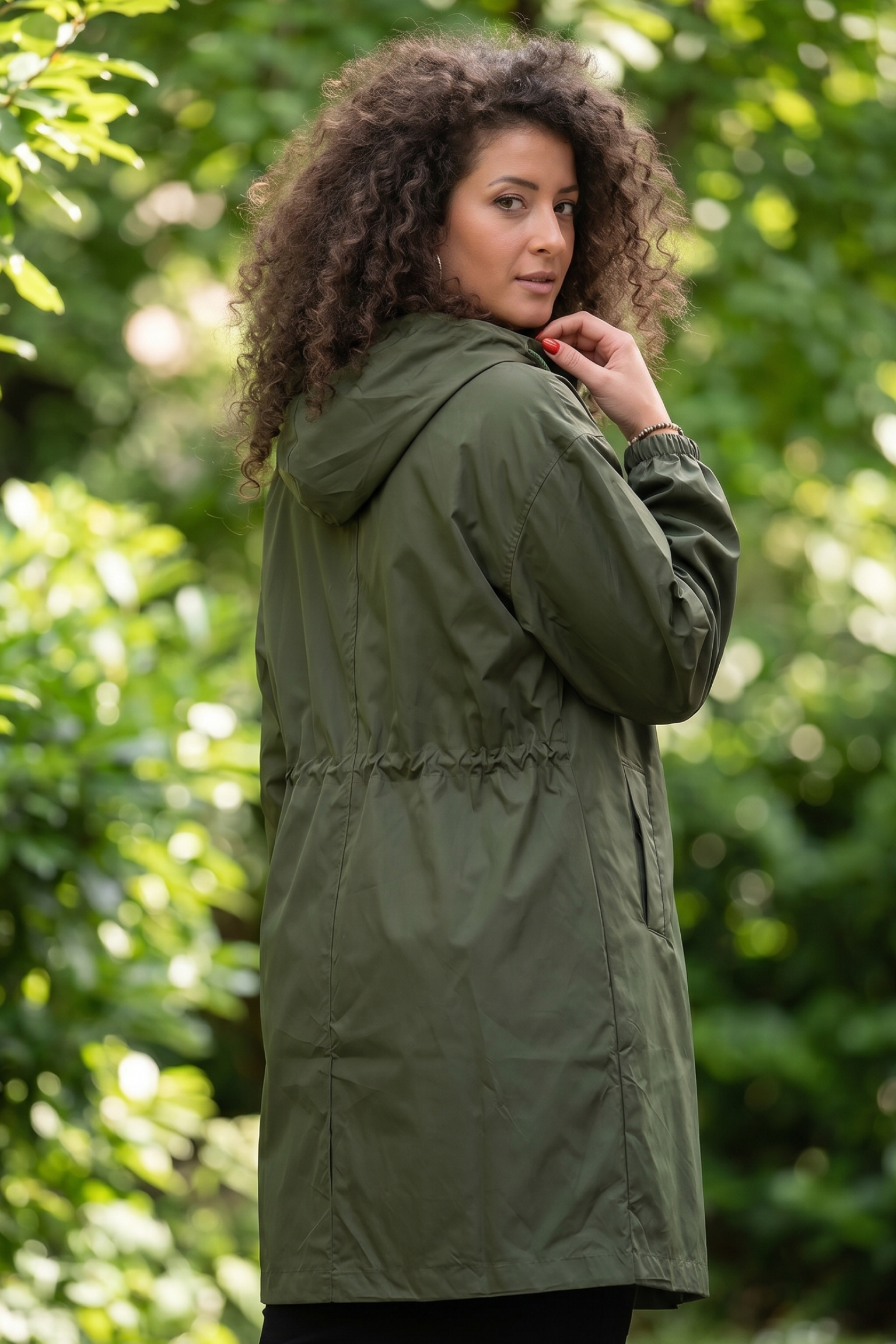 A woman with curly hair wears The Water Resistant Hooded Oversized Windbreaker Rain Jacket in olive green, standing outdoors among sunlit greenery and looking over her shoulder.