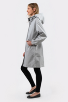 A person stands in profile against a plain white background wearing the Icon Windbreaker Rain Jacket in light gray, paired with black leggings and black flats, with their hands in the jacket’s pockets.
