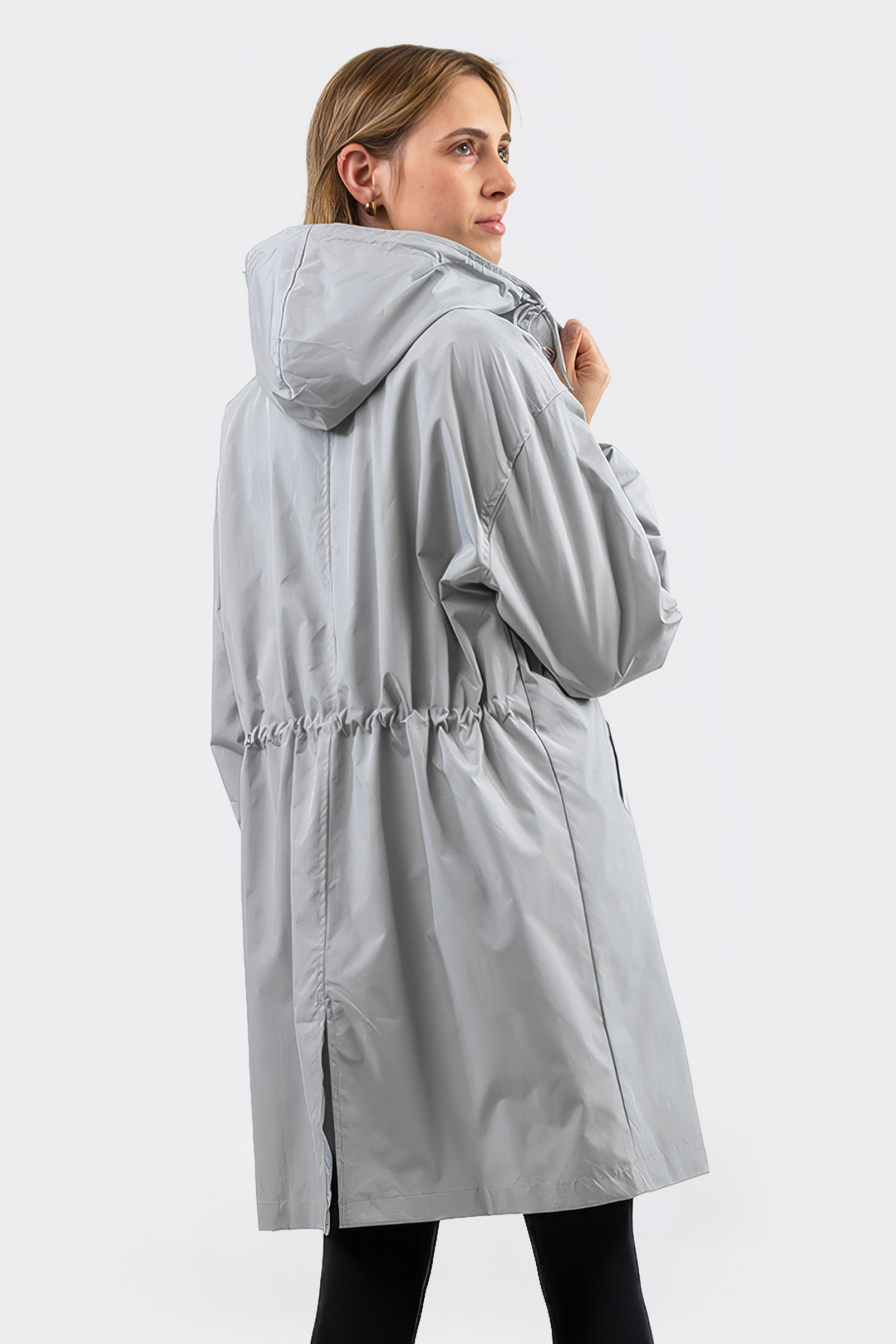A person stands with their back to the camera, head turned right, wearing The Water Resistant Hooded Oversized Windbreaker Rain Jacket in light gray. The loose-fit raincoat features an adjustable waistband against a plain white background.