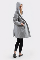 A person models the Icon Windbreaker Rain Jacket in light gray, paired with black leggings and black flats, standing with one hand in the jacket pocket against a plain white background.