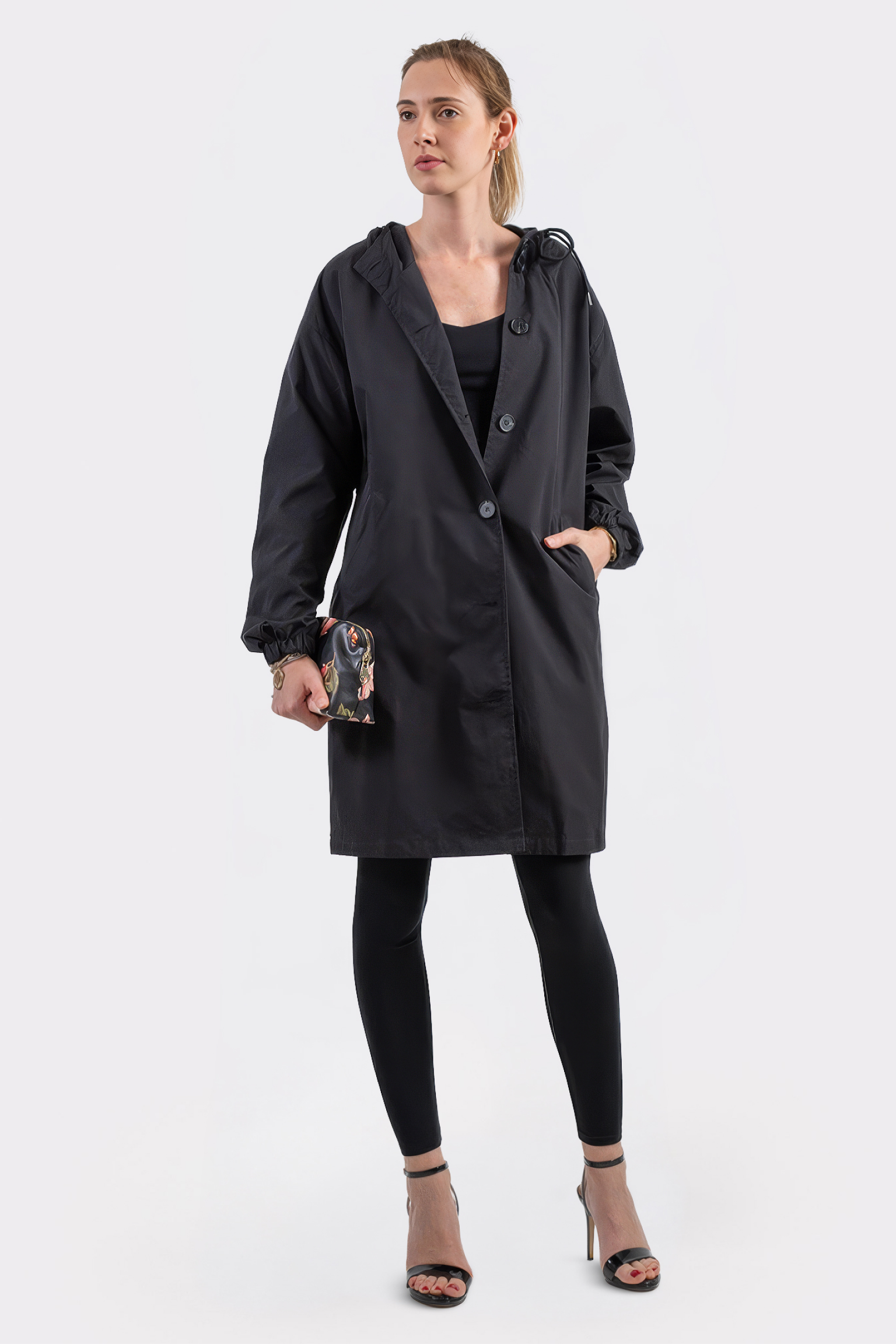 A woman faces forward wearing The Water Resistant Hooded Oversized Windbreaker Rain Jacket in black, paired with black leggings and high-heeled sandals. She holds a small clutch bag, her hair pulled back, against a plain white background.