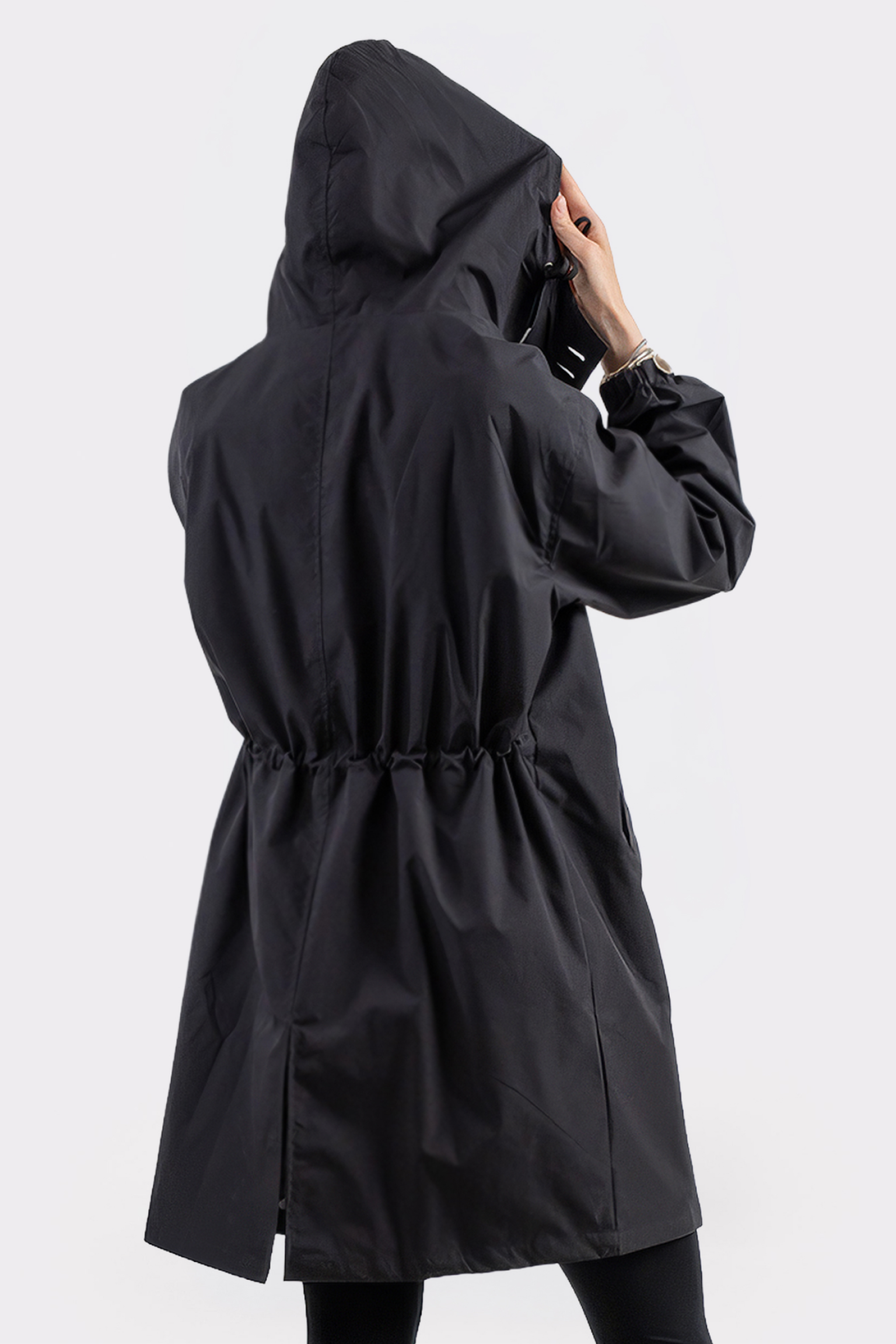 Wearing The Water Resistant Hooded Oversized Windbreaker Rain Jacket, a person stands with their back to the camera, holding the hood. The black jacket features a cinched waist and back hem slit. The background is plain white.