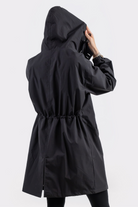 Someone is seen from behind wearing the Icon Windbreaker Rain Jacket—a long black raincoat with a hood, cinched waist, and back hem slit. One hand holds the hood. The background is light and plain.