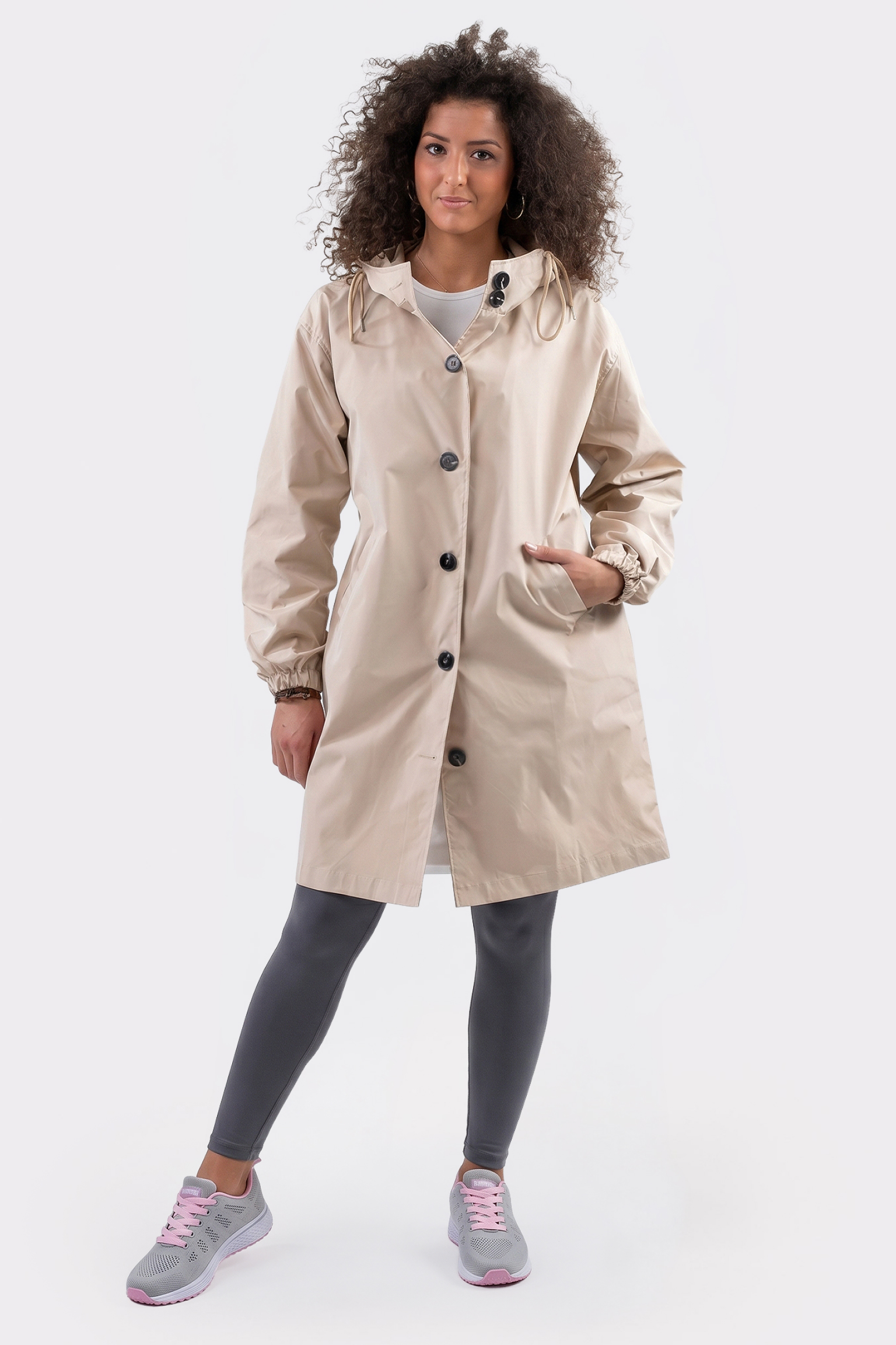 A person stands facing forward in The Water Resistant Hooded Oversized Windbreaker Rain Jacket, paired with gray leggings and gray sneakers with pink accents, against a plain white background.