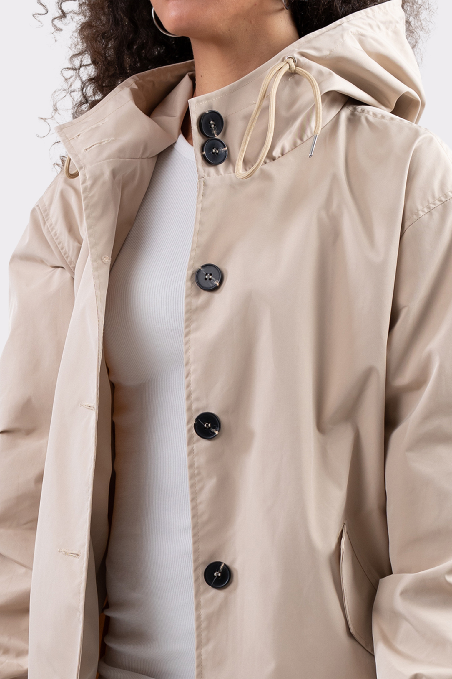 A person wears The Water Resistant Hooded Oversized Windbreaker Rain Jacket in light beige, featuring black buttons, over a white ribbed shirt. Only the lower face and upper torso are shown; background is plain light grey.