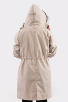 A person is seen from behind wearing the Icon Windbreaker Rain Jacket in light beige, holding the hood with both hands. The jacket features a drawstring waist and long sleeves, set against a plain white background.