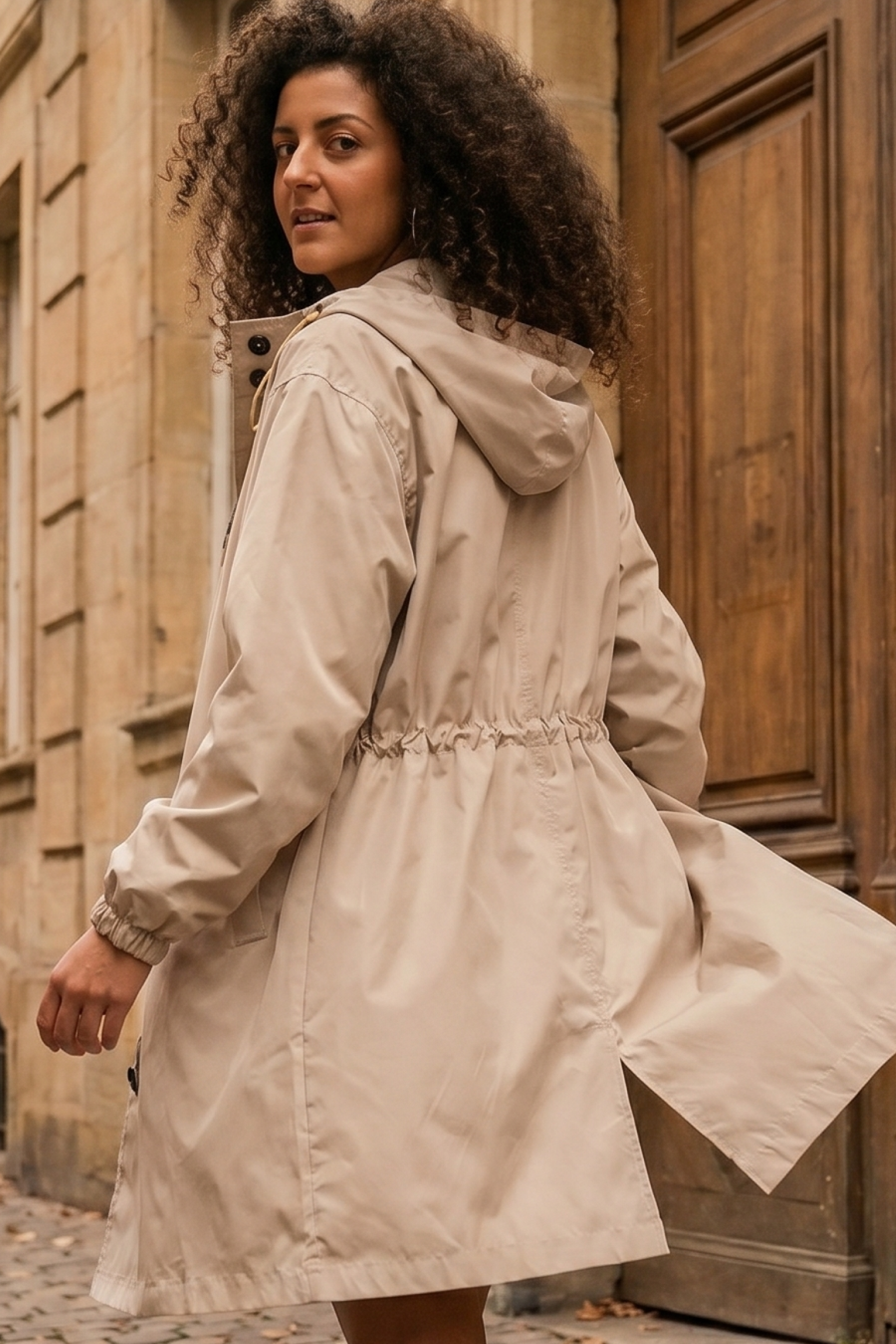 A person with curly hair stands outdoors near a stone building and wooden door, looking back while wearing The Water Resistant Hooded Oversized Windbreaker Rain Jacket in light beige, the coat slightly billowing.