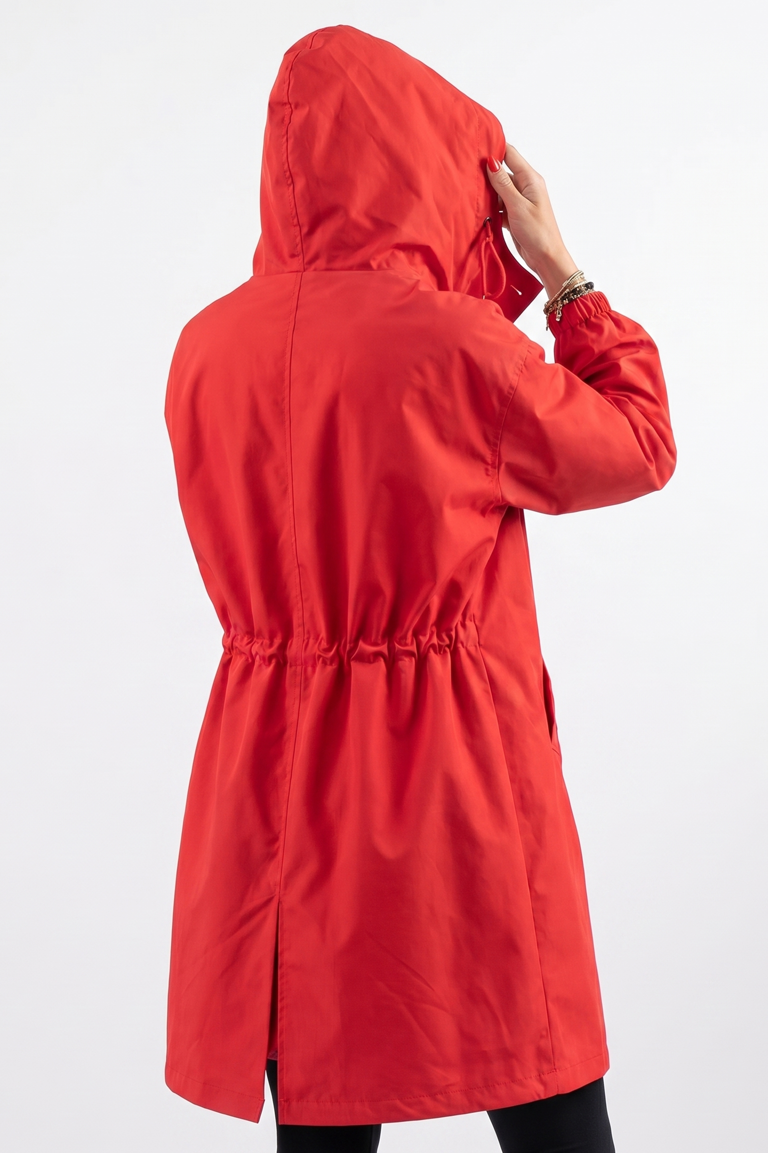 Someone wearing The Water Resistant Hooded Oversized Windbreaker Rain Jacket in bright red stands with their back to the camera, holding the hood in one hand. The jacket features a gathered waist and a slit at the back hem.