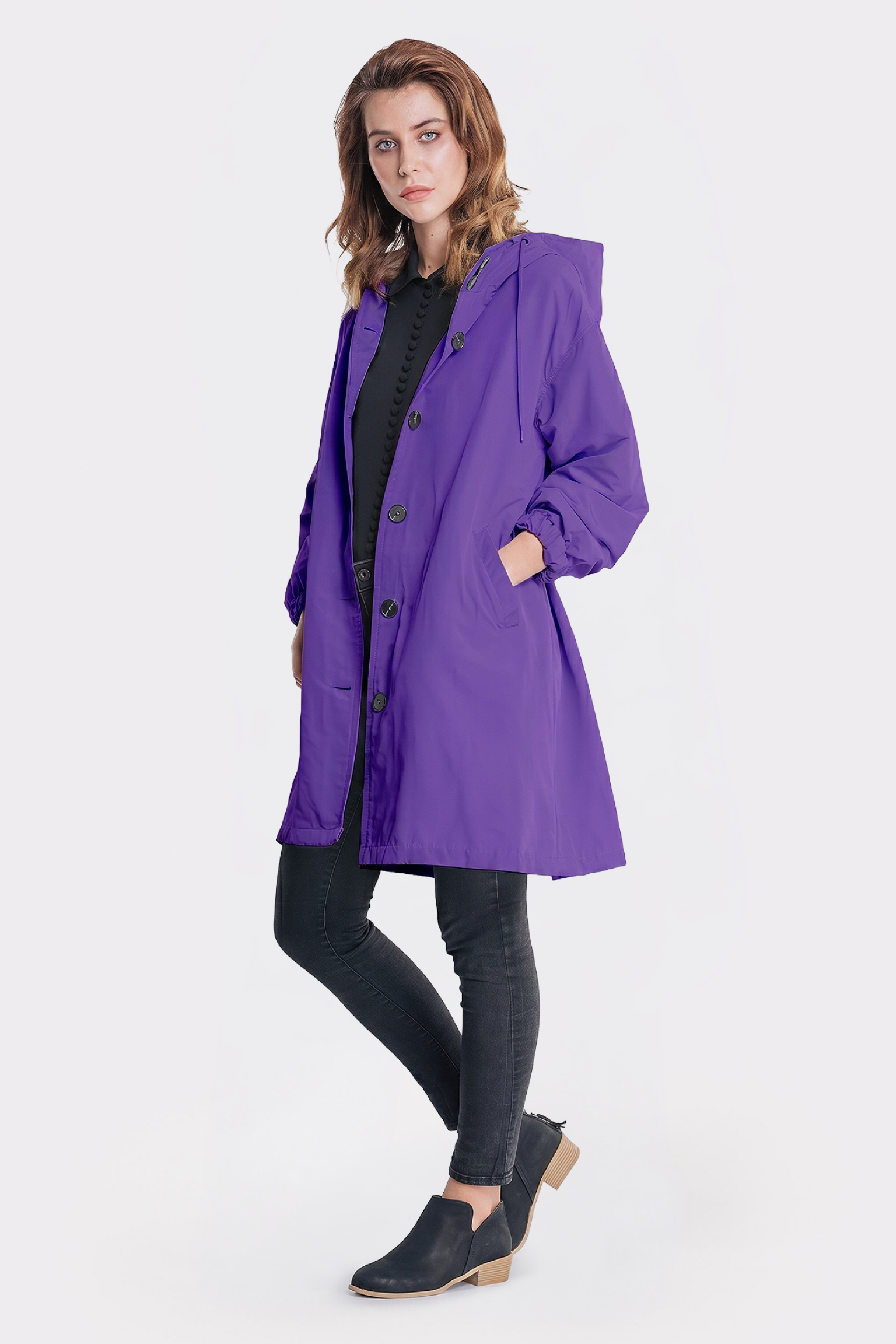 A person stands facing forward in The Water Resistant Hooded Oversized Windbreaker Rain Jacket, a long purple raincoat, over a black shirt, skinny black jeans, and low-heeled black shoes. One hand is in a pocket against a plain white background.