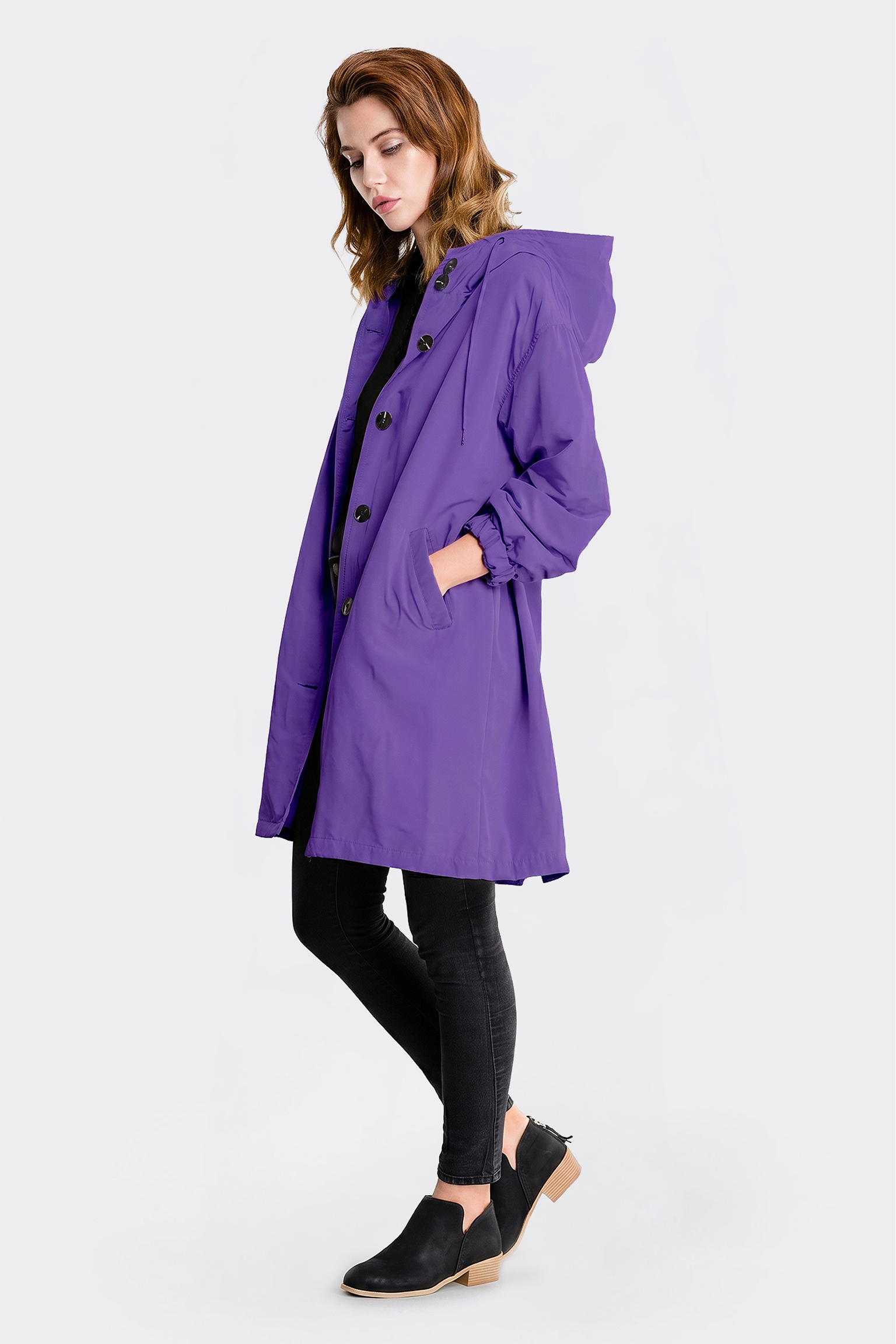 A woman wears The Water Resistant Hooded Oversized Windbreaker Rain Jacket in purple, along with black pants and black slip-on shoes, standing against a plain white background with one hand in her pocket, facing left in a relaxed pose.