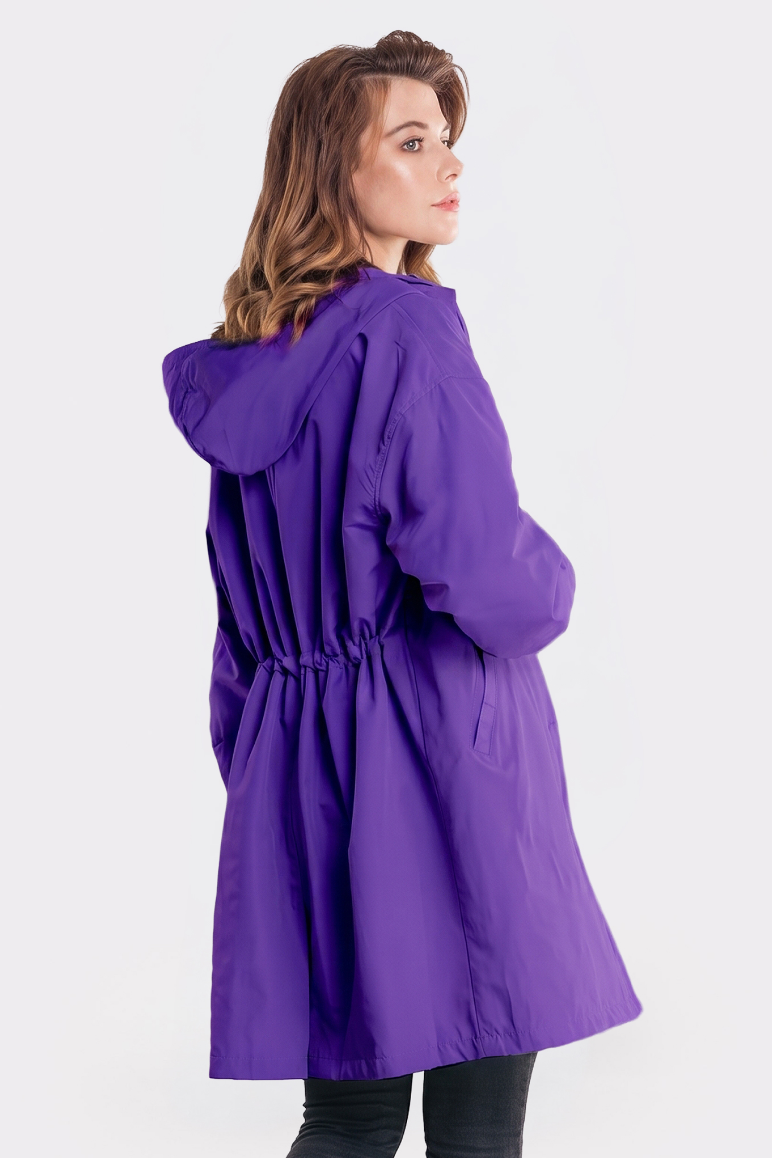 A person in The Water Resistant Hooded Oversized Windbreaker Rain Jacket, featuring long sleeves, a hood, elastic waistband, and side pockets in purple, stands looking over their shoulder against a plain light background.