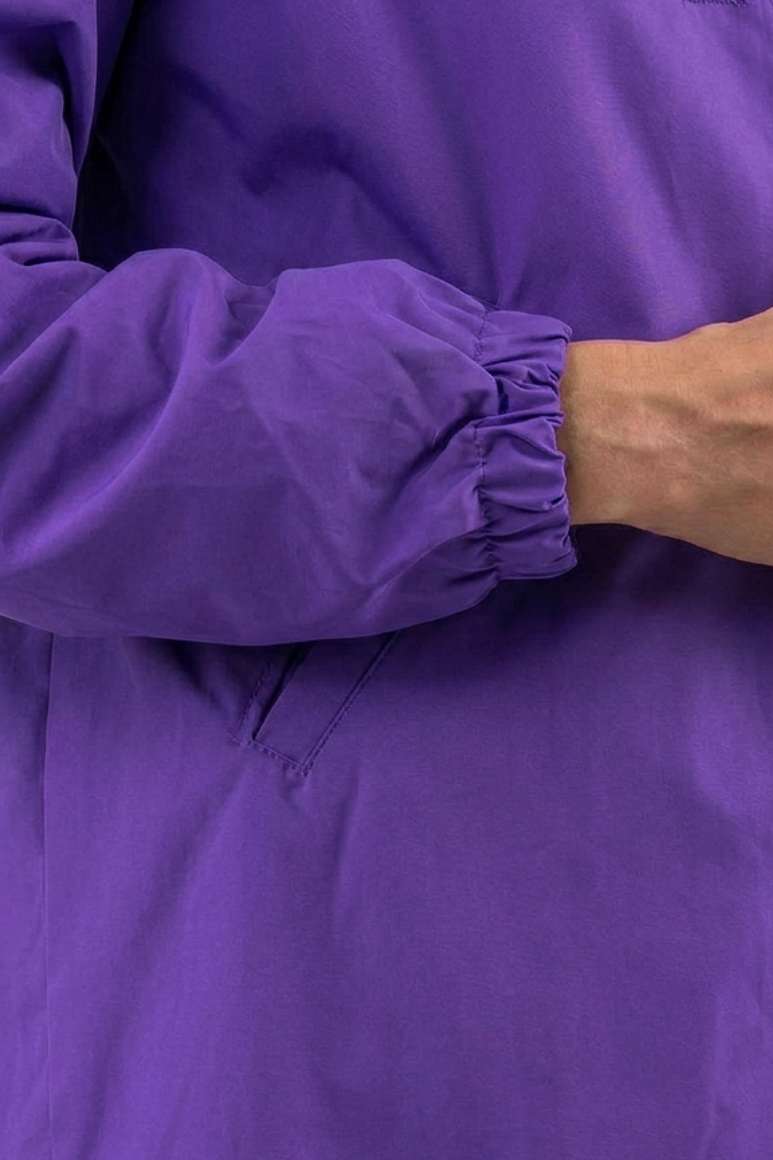 A close-up of a person’s arm and hand wearing The Water Resistant Hooded Oversized Windbreaker Rain Jacket in bright purple with elastic at the wrist, set against a plain background.