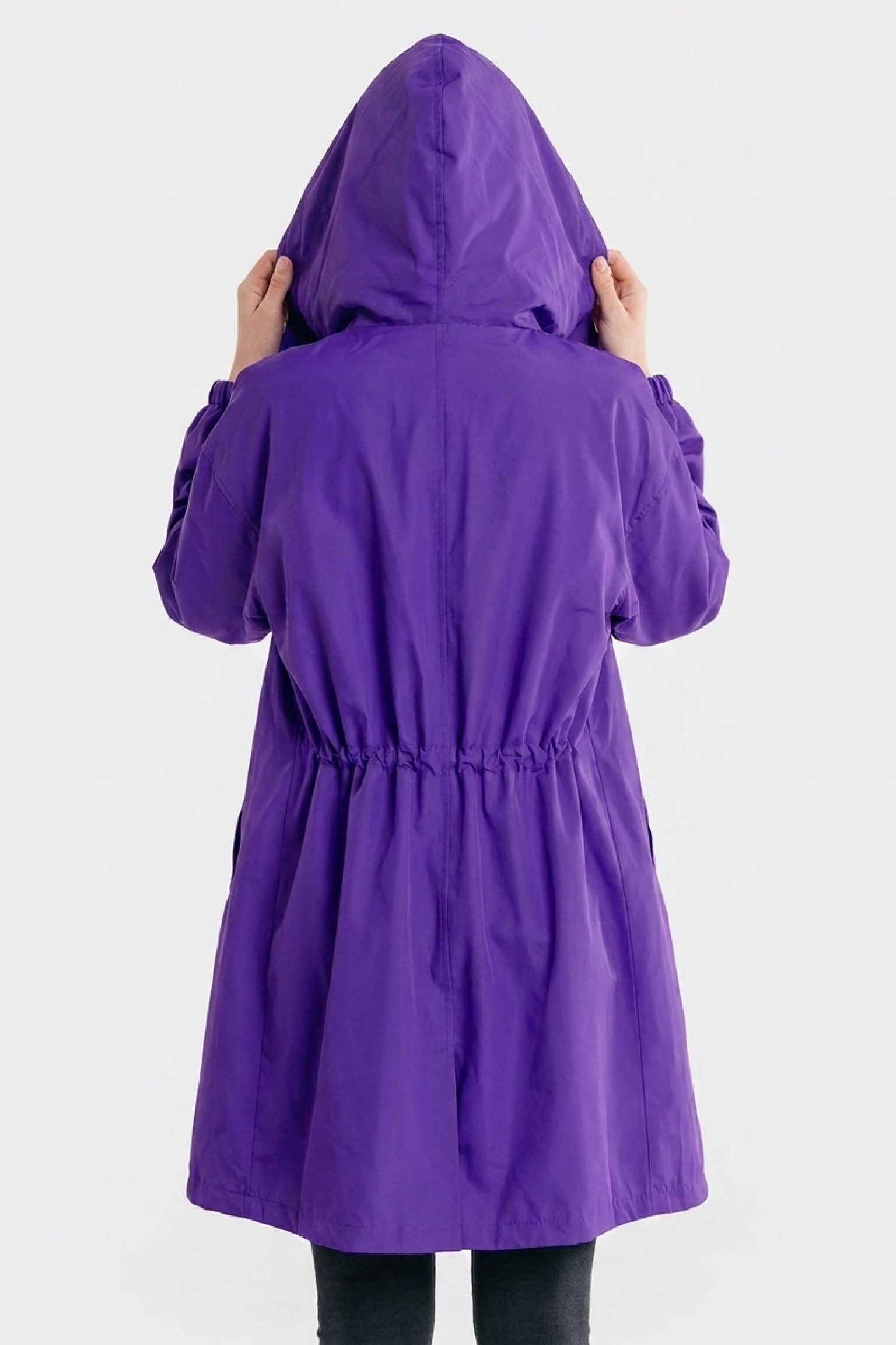 A person with their back to the camera wears the Icon Windbreaker Rain Jacket in purple, hood up and hands holding the sides. The background is plain and light-colored.