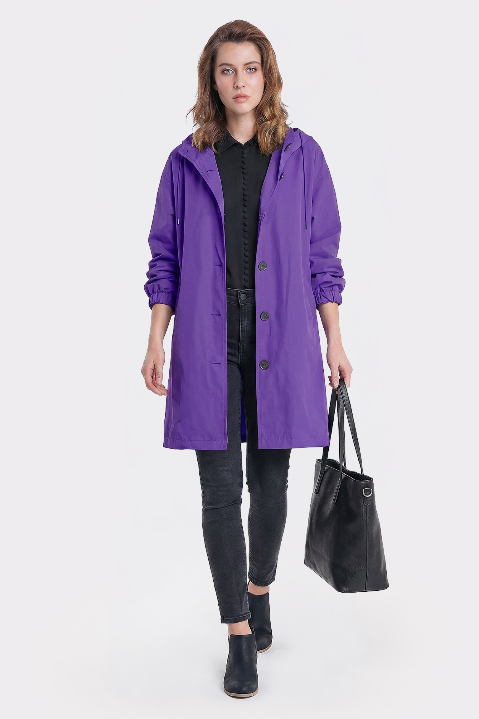 A person models The Water Resistant Hooded Oversized Windbreaker Rain Jacket in purple, layered over a black shirt and pants, carrying a large black tote bag in their left hand against a plain white background.