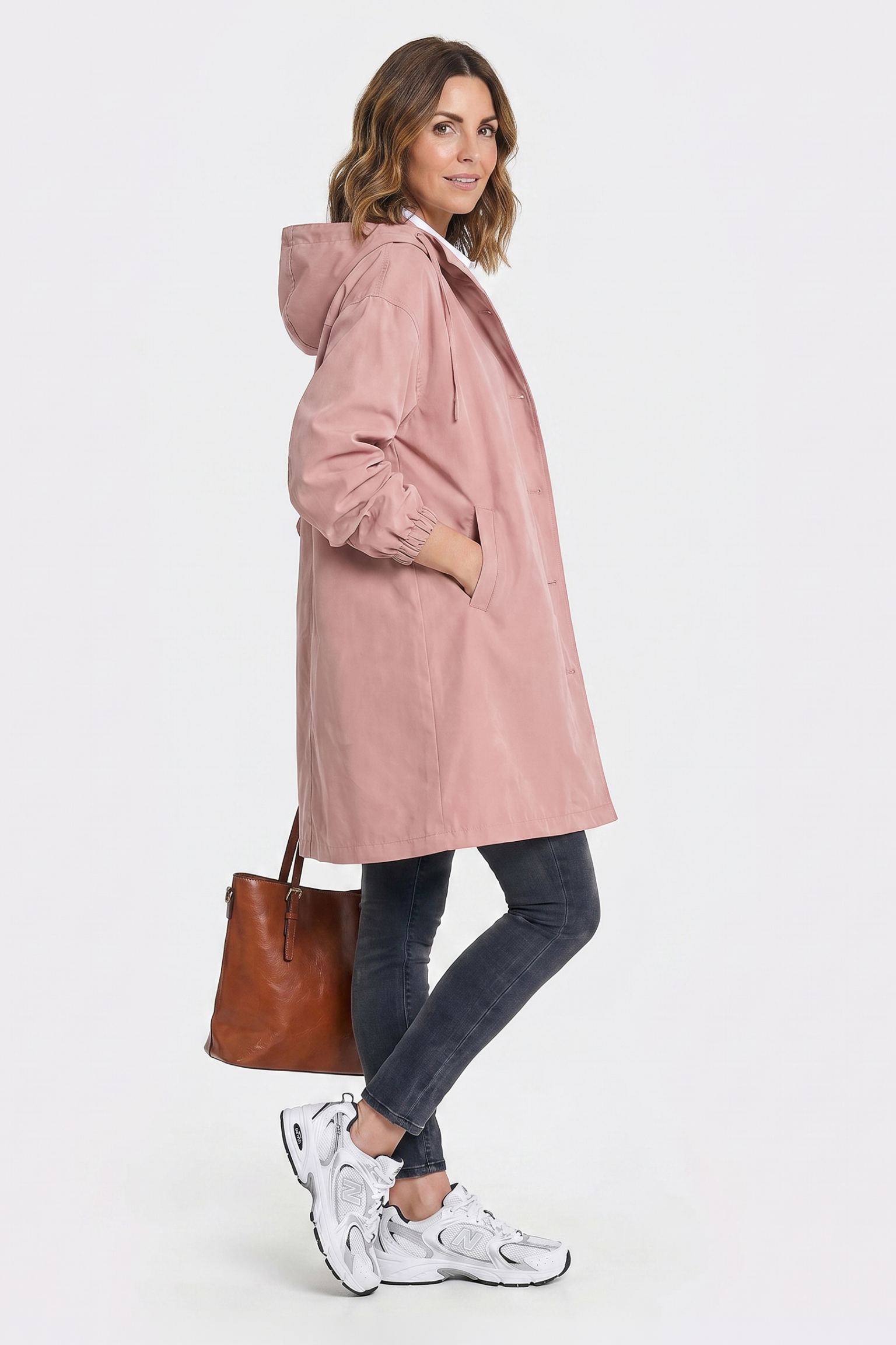 A woman models The Water Resistant Hooded Oversized Windbreaker Rain Jacket, dark jeans, white sneakers, and a brown leather tote bag against a white background.