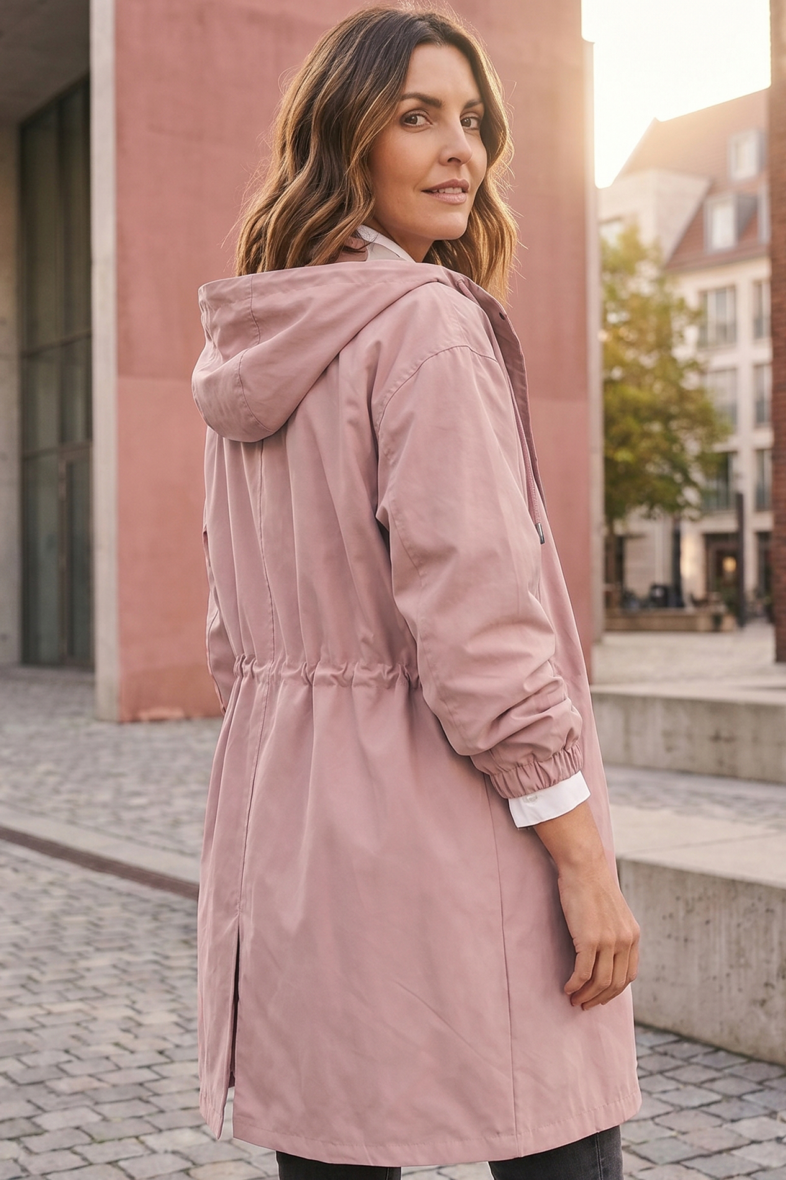 A woman with wavy brown hair stands outdoors, looking over her shoulder, wearing The Water Resistant Hooded Oversized Windbreaker Rain Jacket in light pink. Buildings and trees can be seen in the background.