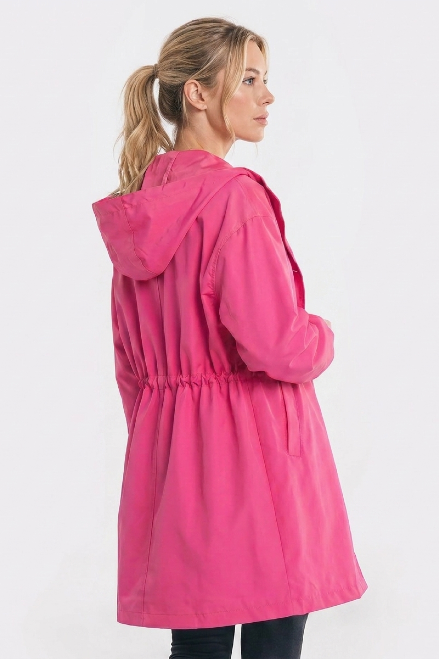 A person with blonde hair in a ponytail models the Icon Windbreaker Rain Jacket, featuring a bright pink color and cinched waist, standing sideways against a plain white background.