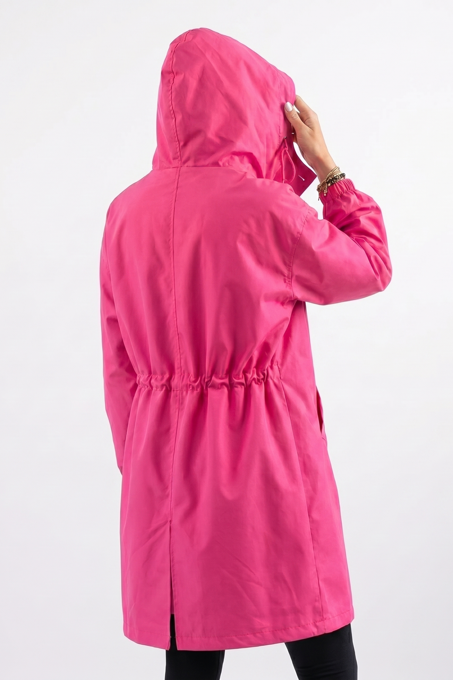 A person wearing The Water Resistant Hooded Oversized Windbreaker Rain Jacket in bright pink is shown from the back, adjusting the hood. The jacket features an elasticized waist against a plain white background.
