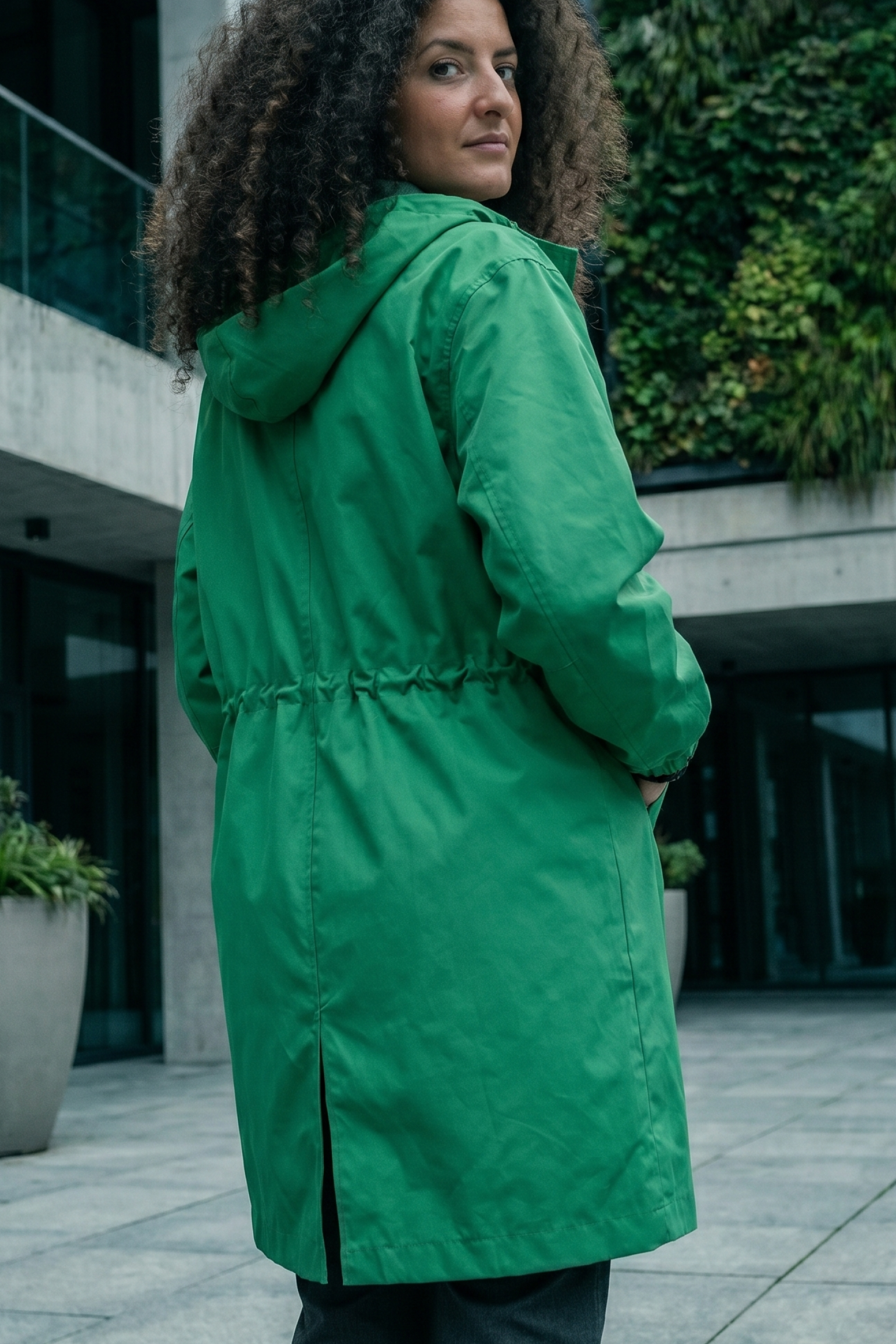 A woman with curly brown hair stands facing away from the camera, looking over her shoulder while wearing the Icon Windbreaker Rain Jacket in bright green.