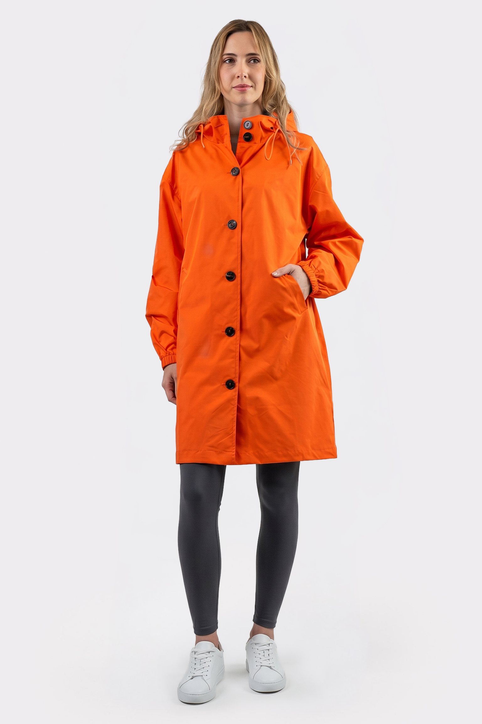A person stands facing forward, wearing The Water Resistant Hooded Oversized Windbreaker Rain Jacket in bright orange, paired with black leggings and white sneakers against a plain light background.