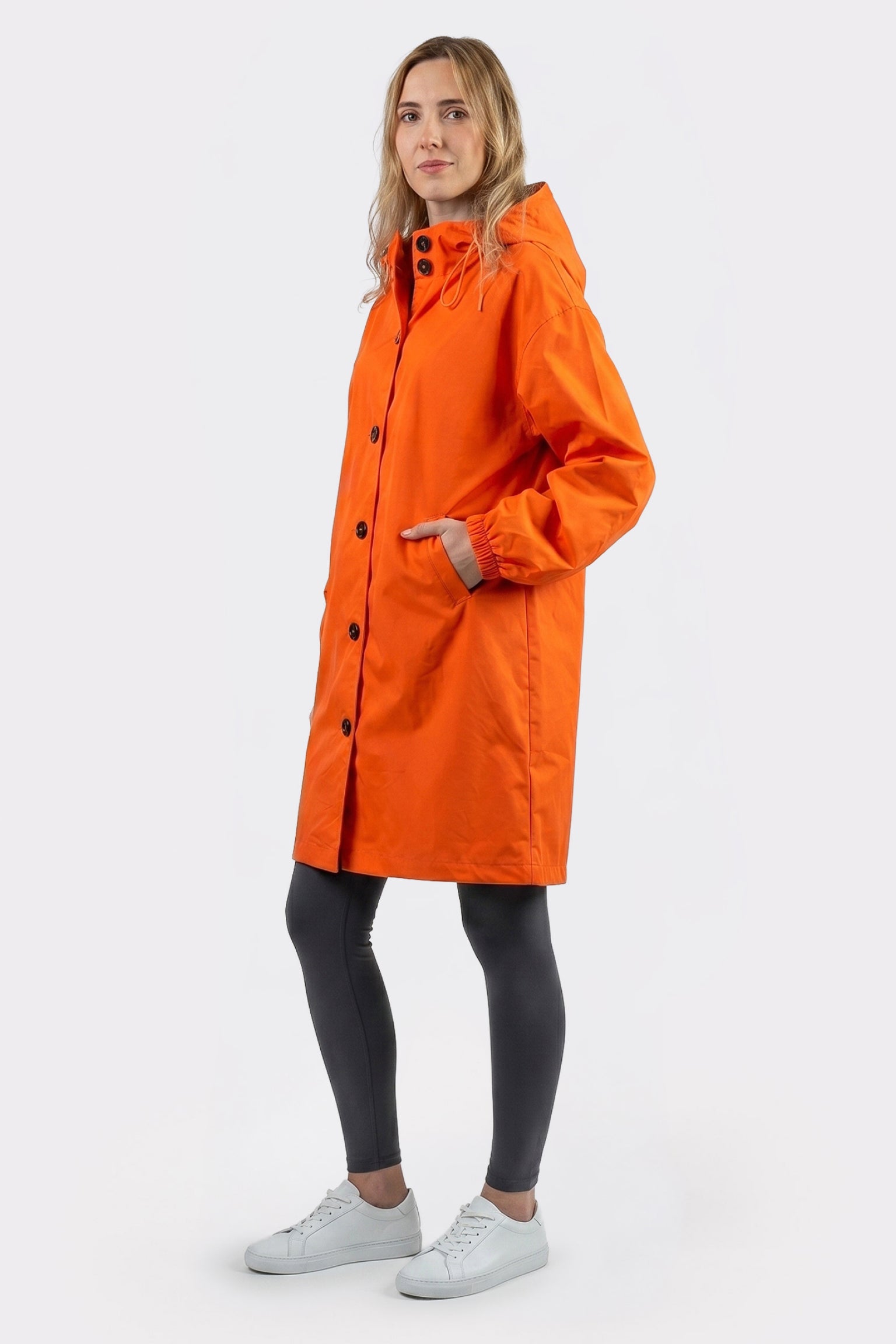 A person stands against a plain background wearing The Water Resistant Hooded Oversized Windbreaker Rain Jacket in orange, paired with dark leggings and white sneakers, one hand in a pocket and a neutral expression.