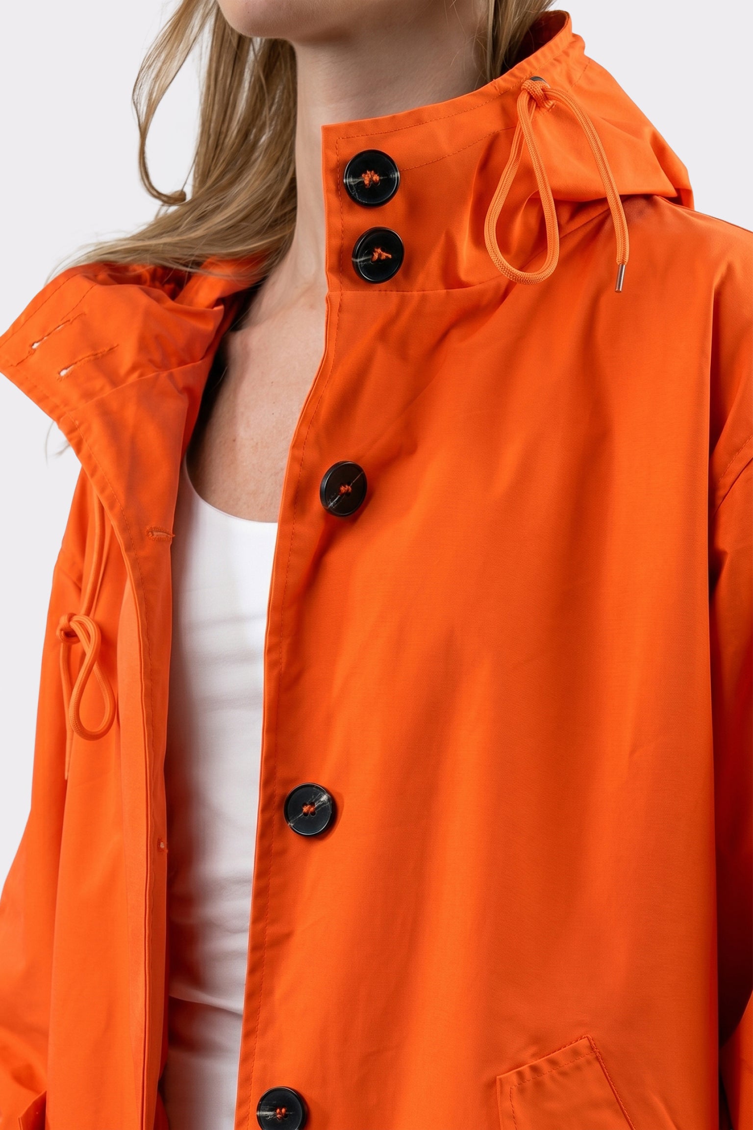 A person models The Water Resistant Hooded Oversized Windbreaker Rain Jacket in orange, featuring large black buttons over a white top. The cropped photo highlights the jacket's unique details and upper body fit.