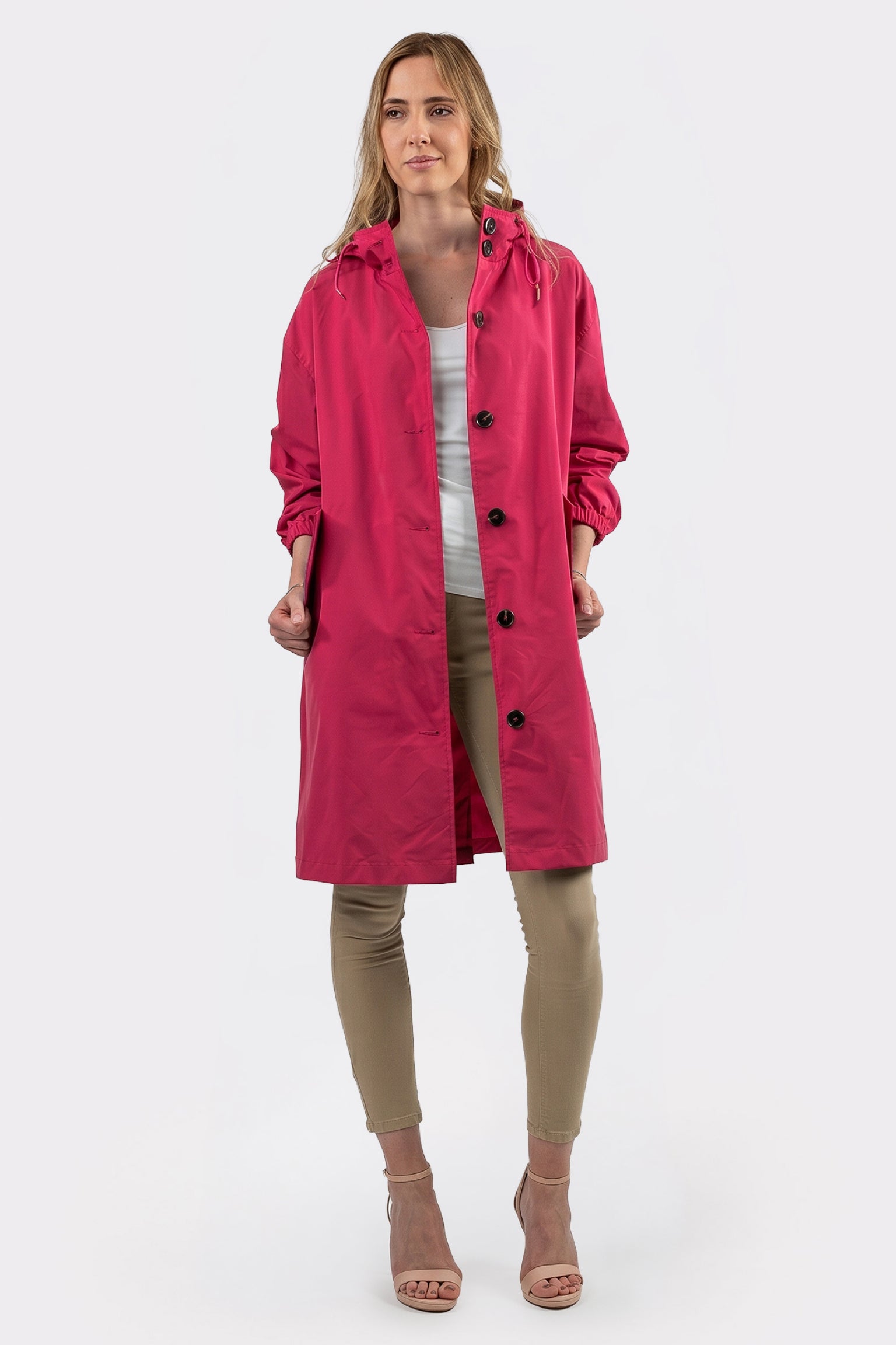 A woman faces forward against a plain white background, wearing The Water Resistant Hooded Oversized Windbreaker Rain Jacket in bright pink over a white top, beige pants, and open-toe heels.
