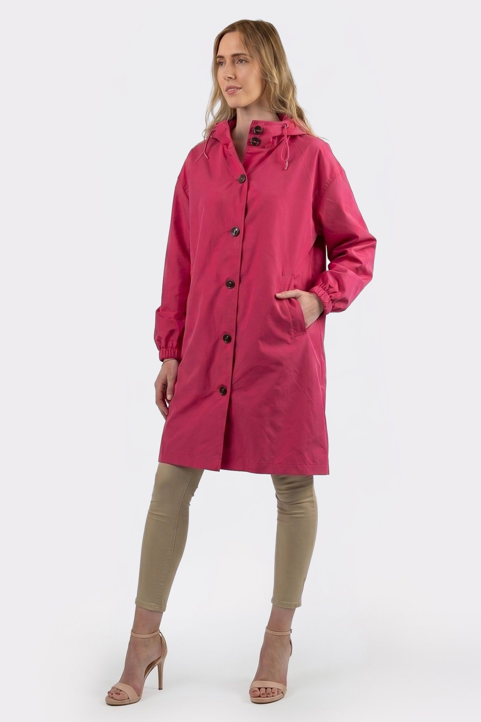 A woman stands against a plain white background wearing The Water Resistant Hooded Oversized Windbreaker Rain Jacket in bright pink, paired with tan leggings and beige high-heeled sandals.