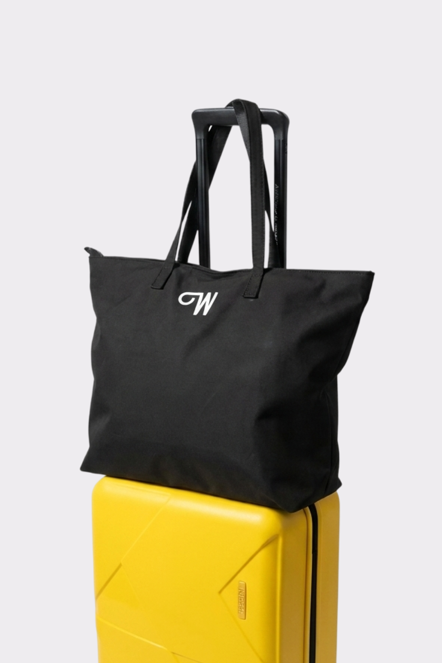 The Urban Slate Waterproof Bag, a spacious black duffel with handles and a white "CW" logo, sits atop a closed yellow hard-shell suitcase with an extended handle against a plain light background.