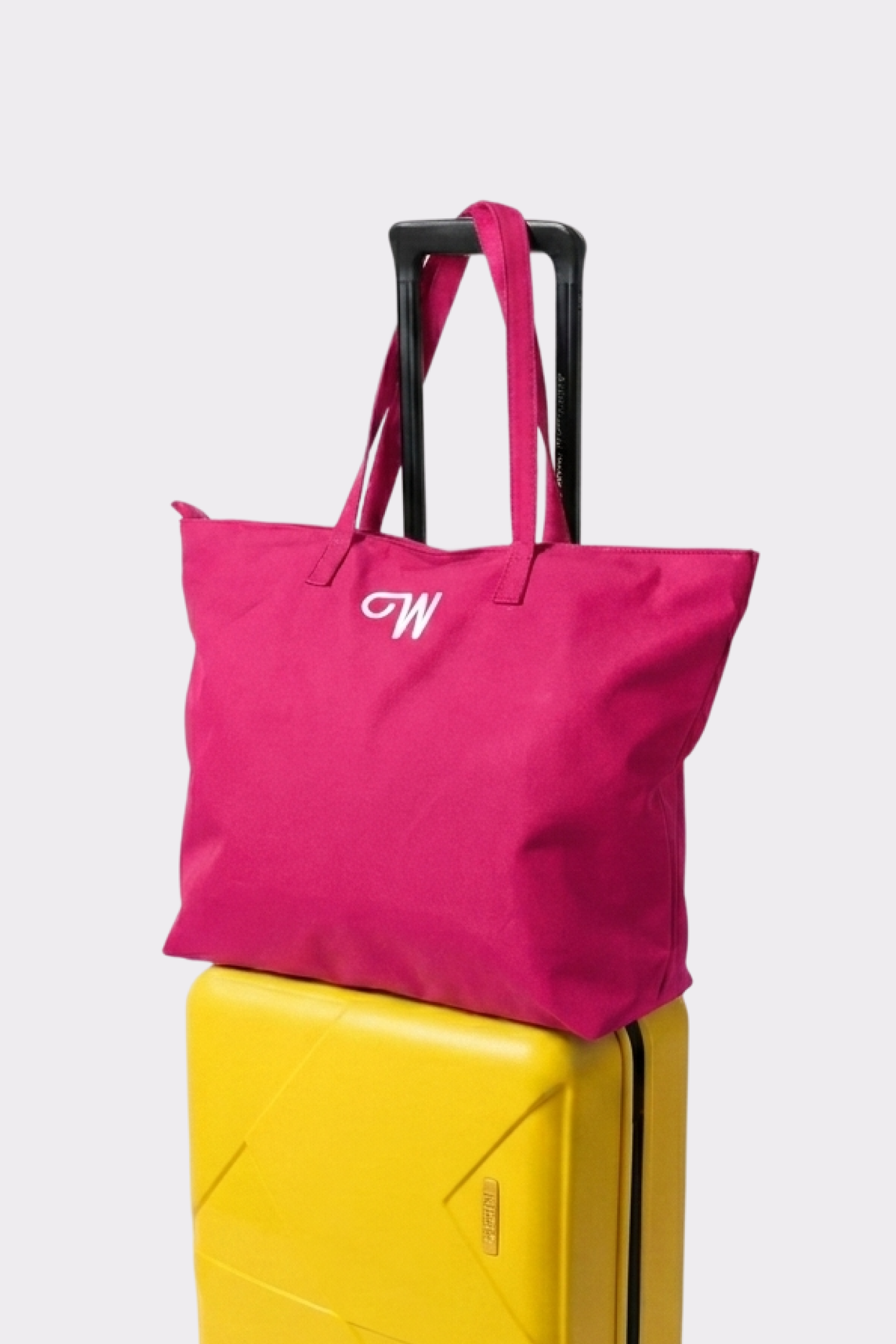The Urban Slate Waterproof Bag, a bright pink travel duffel with two handles and white embroidered initials, sits atop a yellow hard-shell suitcase for stylish organization against a simple light backdrop.